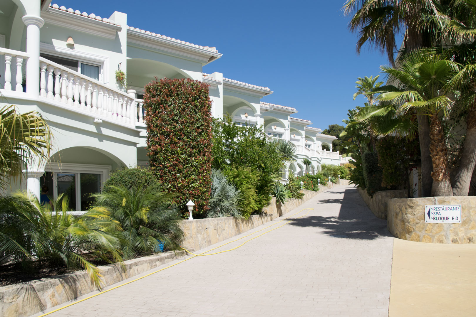 Vista exterior del complejo con fachadas de estilo mediterráneo, terrazas con balaustradas blancas y jardines cuidados junto a un camino pavimentado bajo el sol.