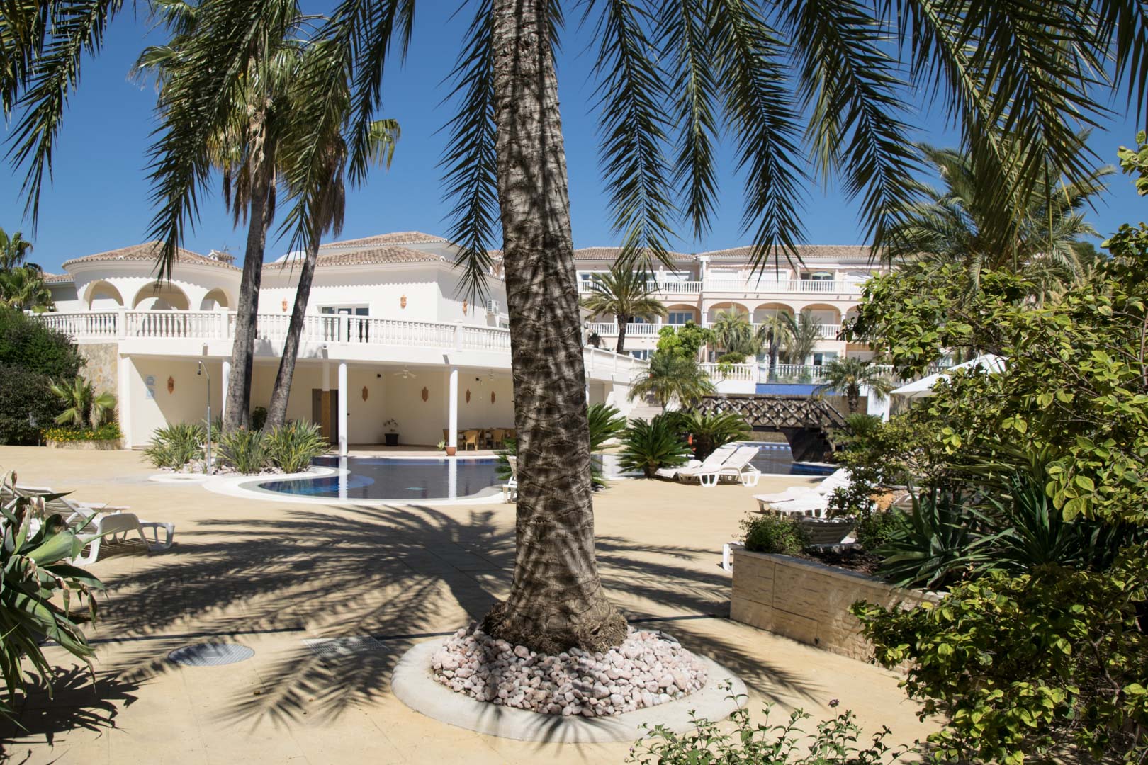 Zona exterior con piscina tipo laguna, rodeada de jardines mediterráneos y palmeras. La arquitectura destaca por sus fachadas blancas, arcos y amplias terrazas soleadas.
