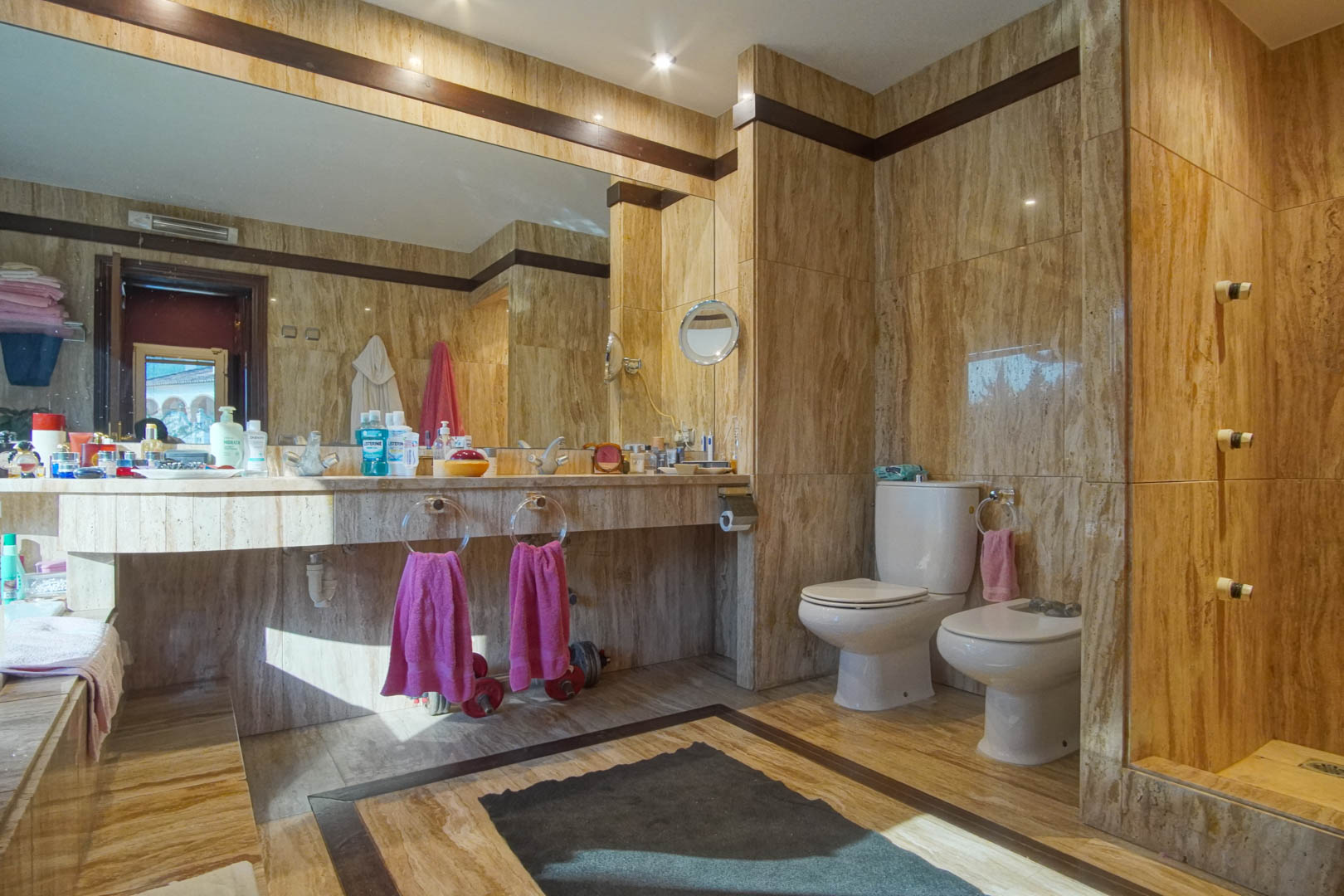 Baño revestido íntegramente en mármol travertino con encimera integrada, gran espejo frontal, sanitarios de porcelana y detalles en madera oscura.
