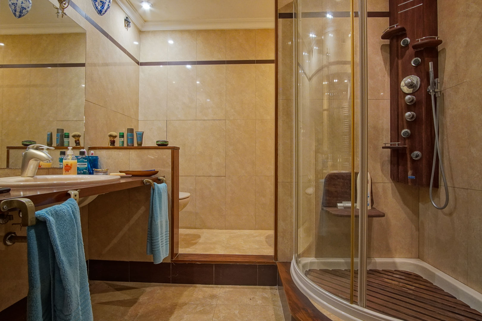 Cuarto de baño con revestimiento de mármol beige, encimera de madera y cabina de ducha curva con sistema de hidromasaje y suelo de lamas de madera.