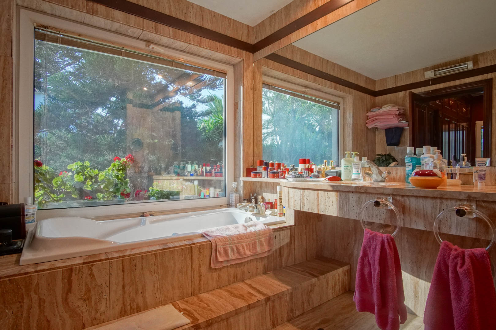 Cuarto de baño revestido en mármol travertino con bañera encastrada junto a un gran ventanal con vistas al jardín y encimera integrada con amplio espejo.