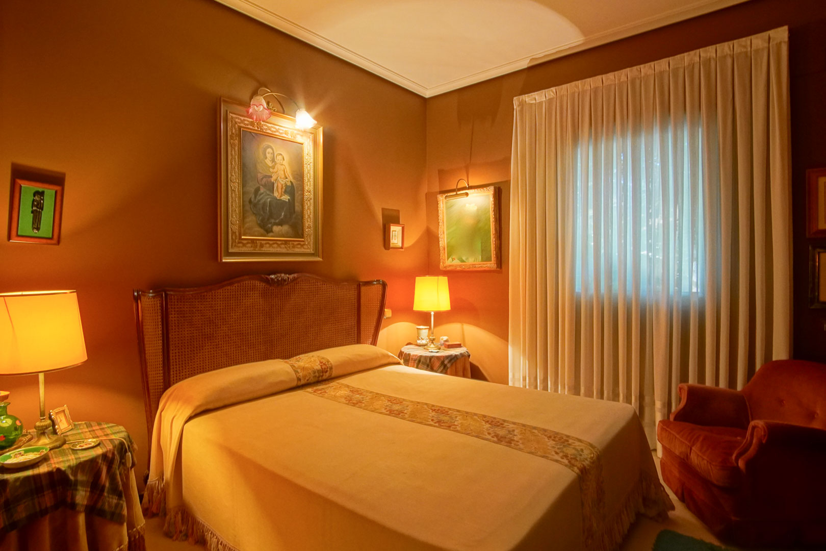 Dormitorio de estilo clásico con paredes en tono terracota, cabecero de rejilla y luz cálida ambiental. Incluye obras de arte iluminadas y un sillón de terciopelo rojo.