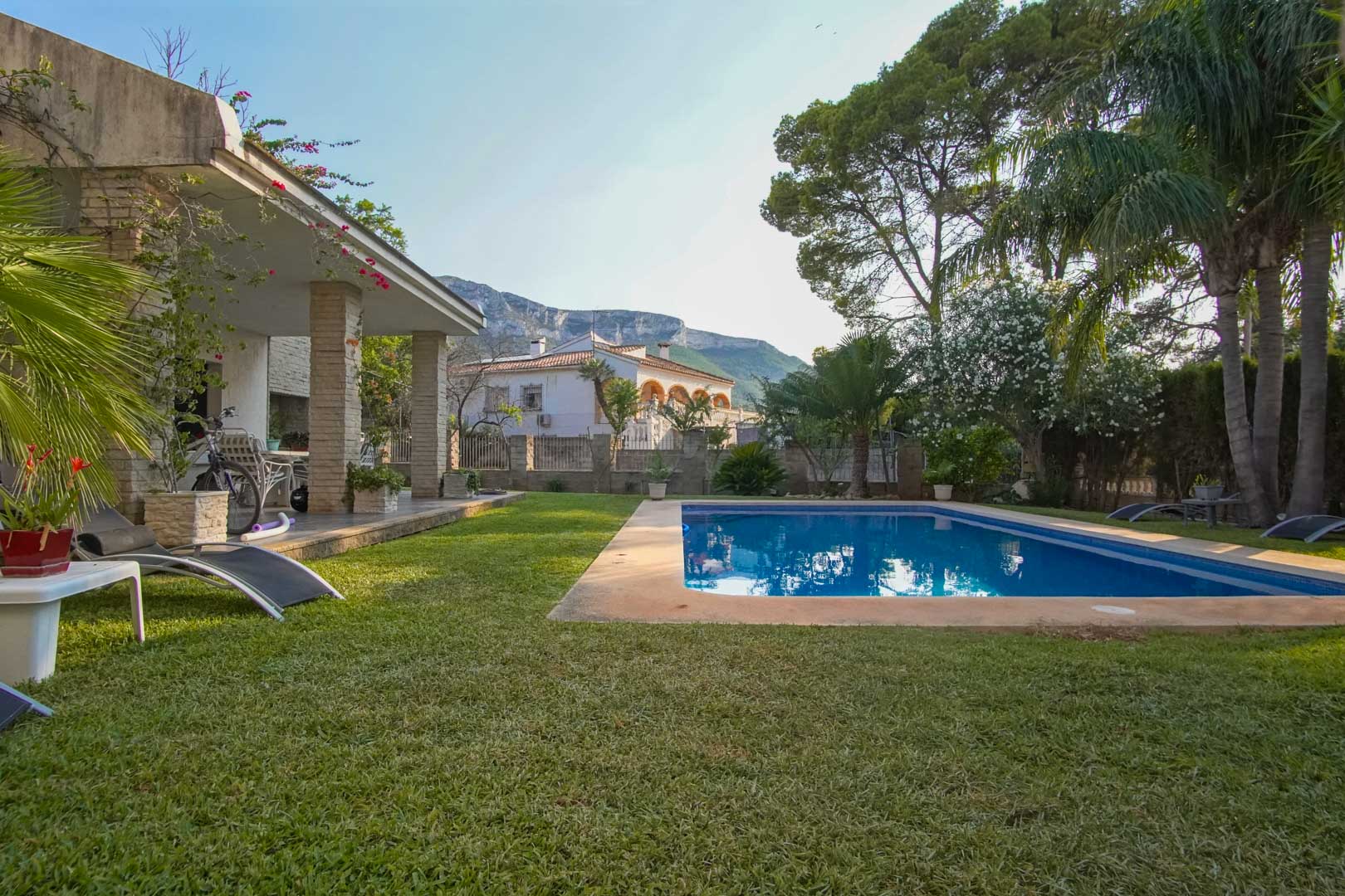 Jardín mediterráneo con césped natural, piscina rectangular y porche con columnas de piedra. Al fondo, destaca la silueta del macizo del Montgó bajo un cielo despejado.