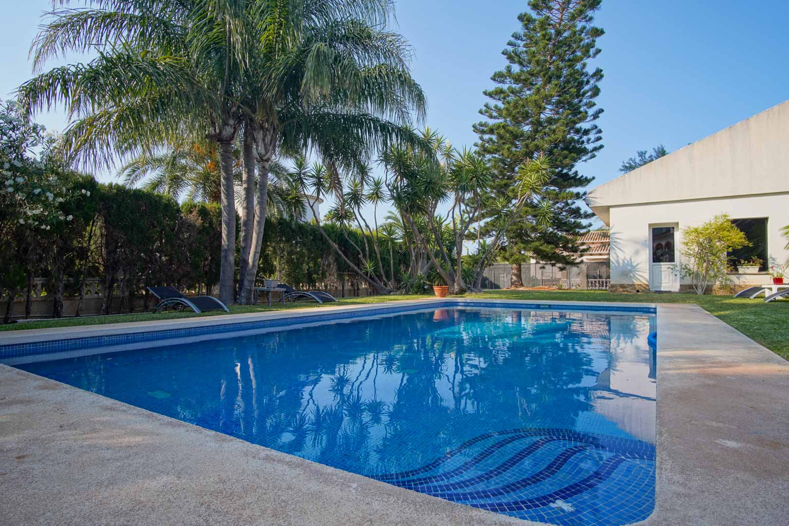 Piscina privada con gresite azul y escalera integrada, rodeada de un jardín mediterráneo con palmeras, césped natural y setos perimetrales que garantizan privacidad.