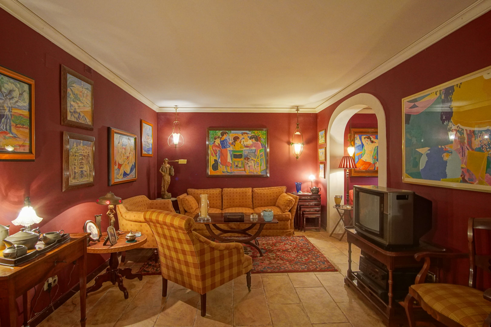 Salón con paredes en tono granate, suelo de gres rústico y mobiliario clásico. Destacan las obras de arte, la iluminación cálida y el acceso mediante un arco decorativo.