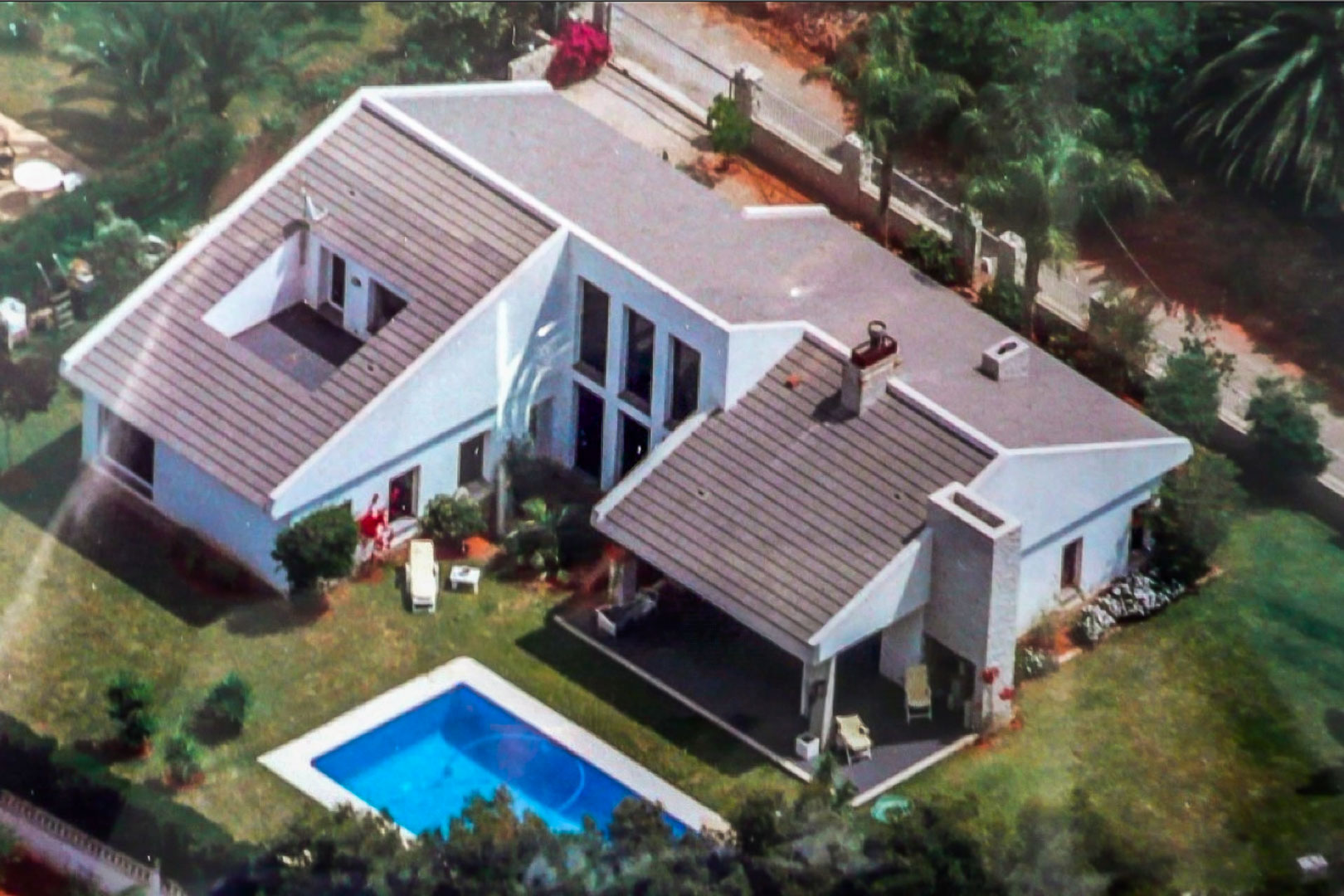 Vista aérea de la villa con fachada blanca, tejado de teja, piscina rectangular y jardín con césped, rodeada de vegetación mediterránea y muros de privacidad.