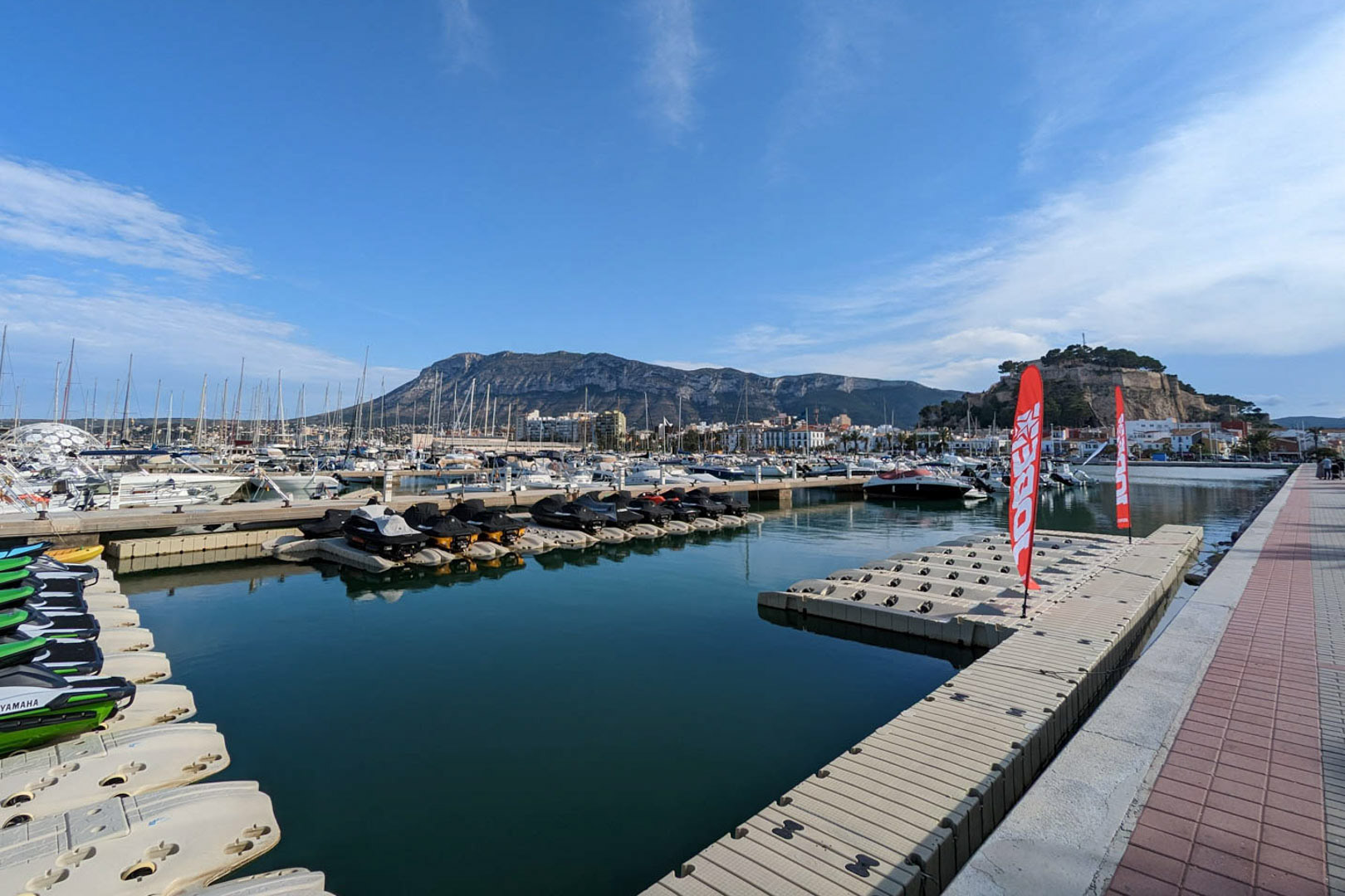 Vista panorámica del puerto deportivo de Dénia con el castillo histórico y el macizo del Montgó al fondo, destacando los amarres y motos de agua.