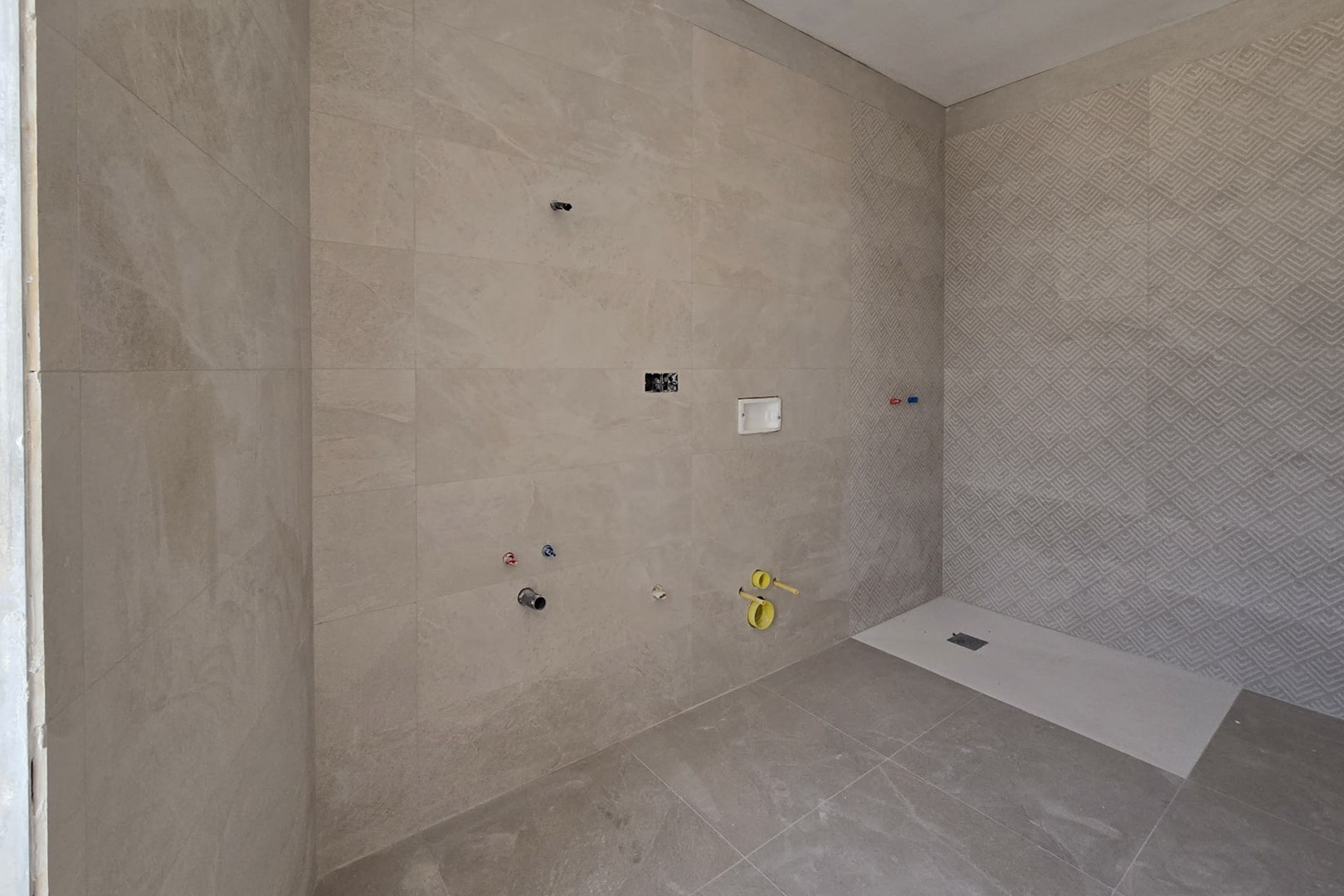 Baño en construcción con revestimiento cerámico en tonos arena, pared de ducha con relieve geométrico y plato de ducha de resina blanca extraplano.