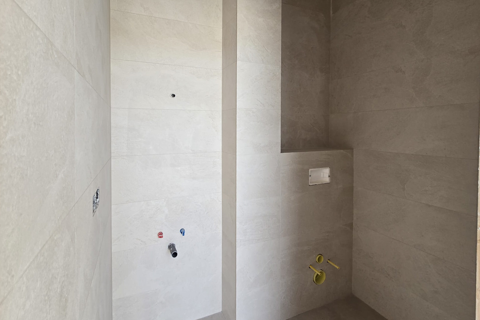 Baño en construcción con revestimiento de gres porcelánico en tonos arena, tomas de agua para lavabo y preinstalación para inodoro suspendido con hornacina integrada.