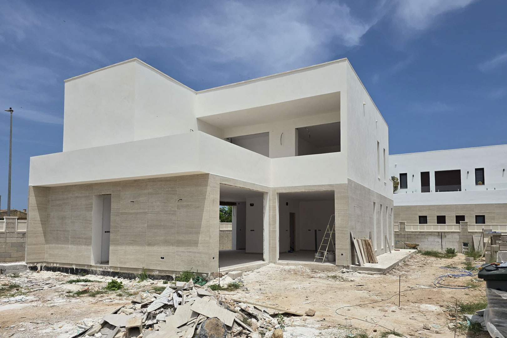 Fachada exterior de villa moderna en construcción con revestimiento de piedra en planta baja, volúmenes geométricos blancos y amplios huecos para ventanales bajo un cielo despejado.