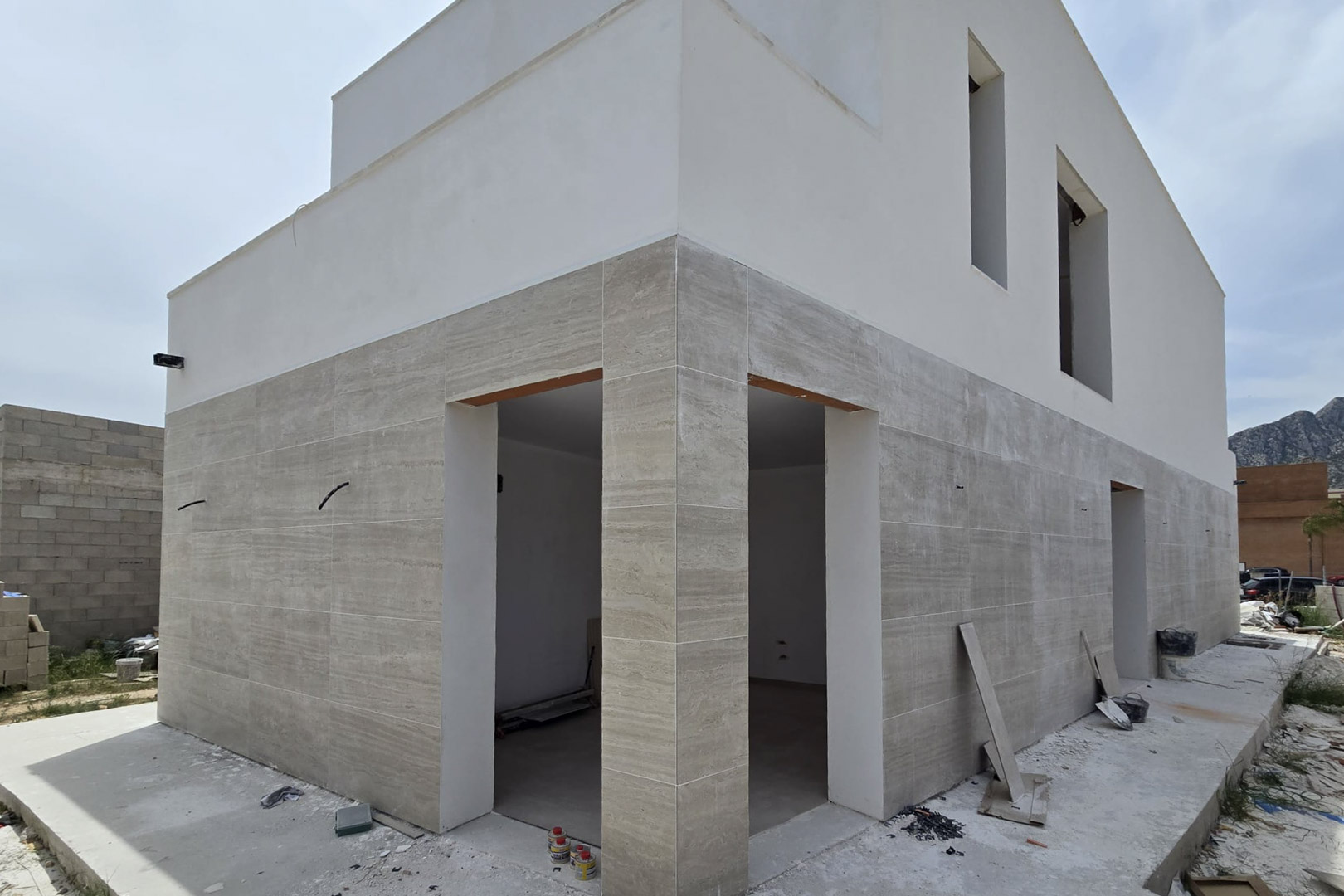 Fachada exterior en construcción con revestimiento de piedra en planta baja y acabado blanco superior, destacando las líneas rectas y amplios huecos para ventanales.