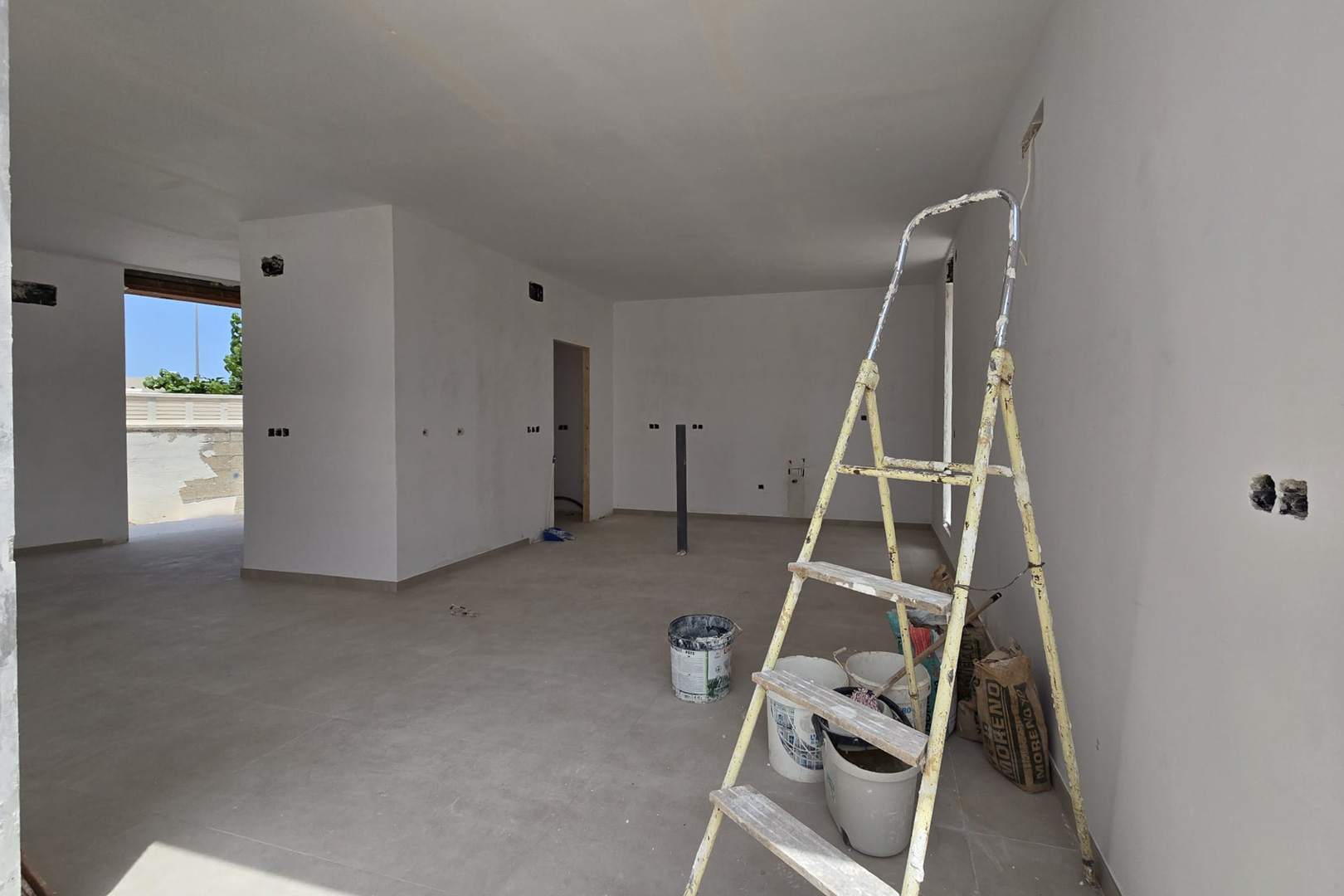 Interior diáfano en fase de acabado con suelos cerámicos de gran formato y paredes blancas. Se aprecia la preinstalación eléctrica y la conexión con la zona exterior.