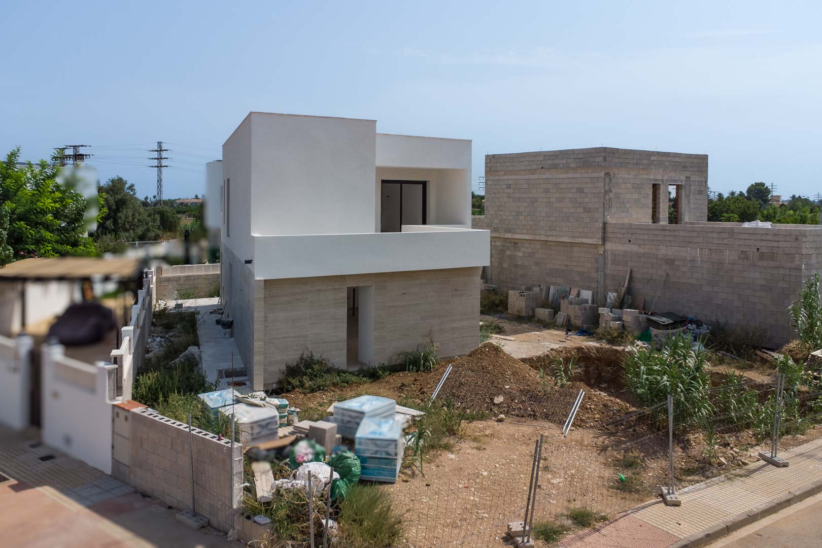 Vista exterior de la villa en construcción con fachada de piedra en planta baja y revoco blanco superior. Se aprecia el hueco excavado para la futura piscina.