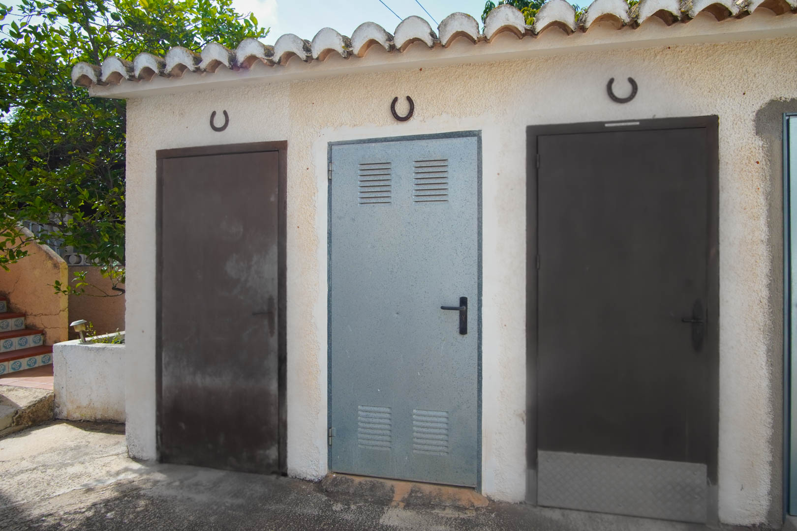 Área de almacenamiento exterior con tres puertas metálicas bajo tejado de teja tradicional, decoradas con herraduras y paredes de acabado rugoso.