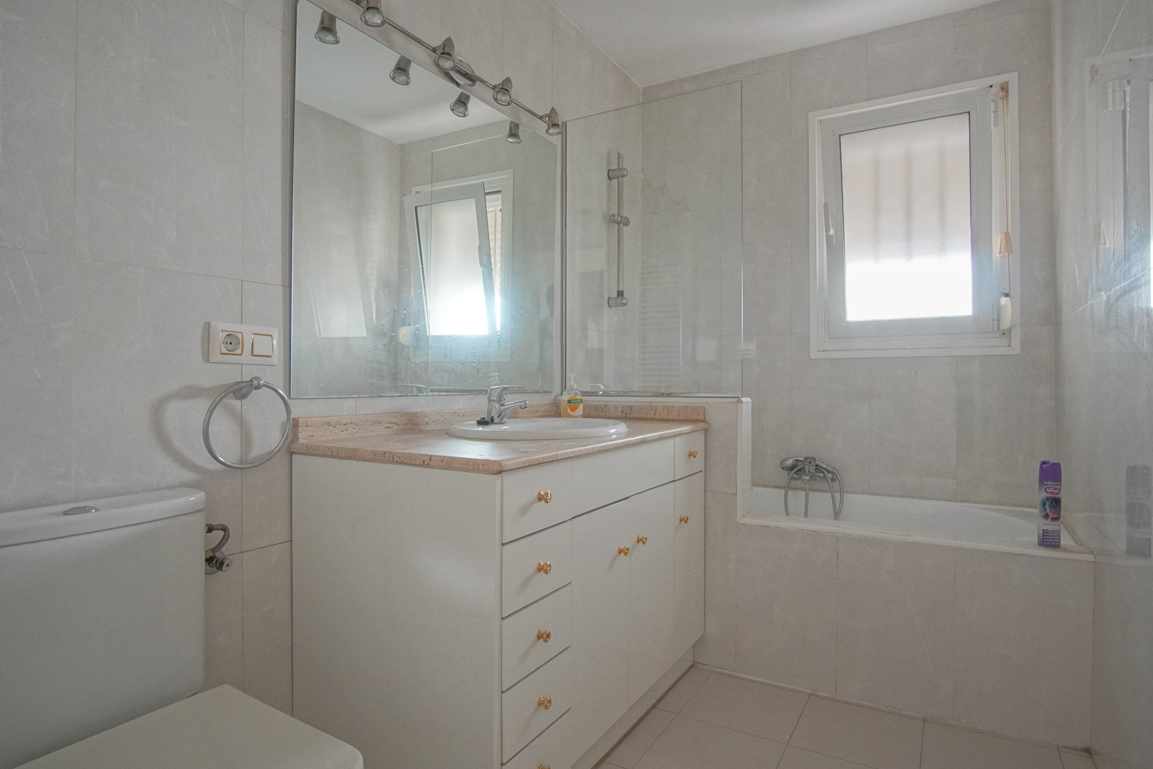 Baño completo con mueble lavabo blanco, encimera de mármol y bañera integrada con mampara de cristal. Dispone de luz natural y ventilación mediante ventana.