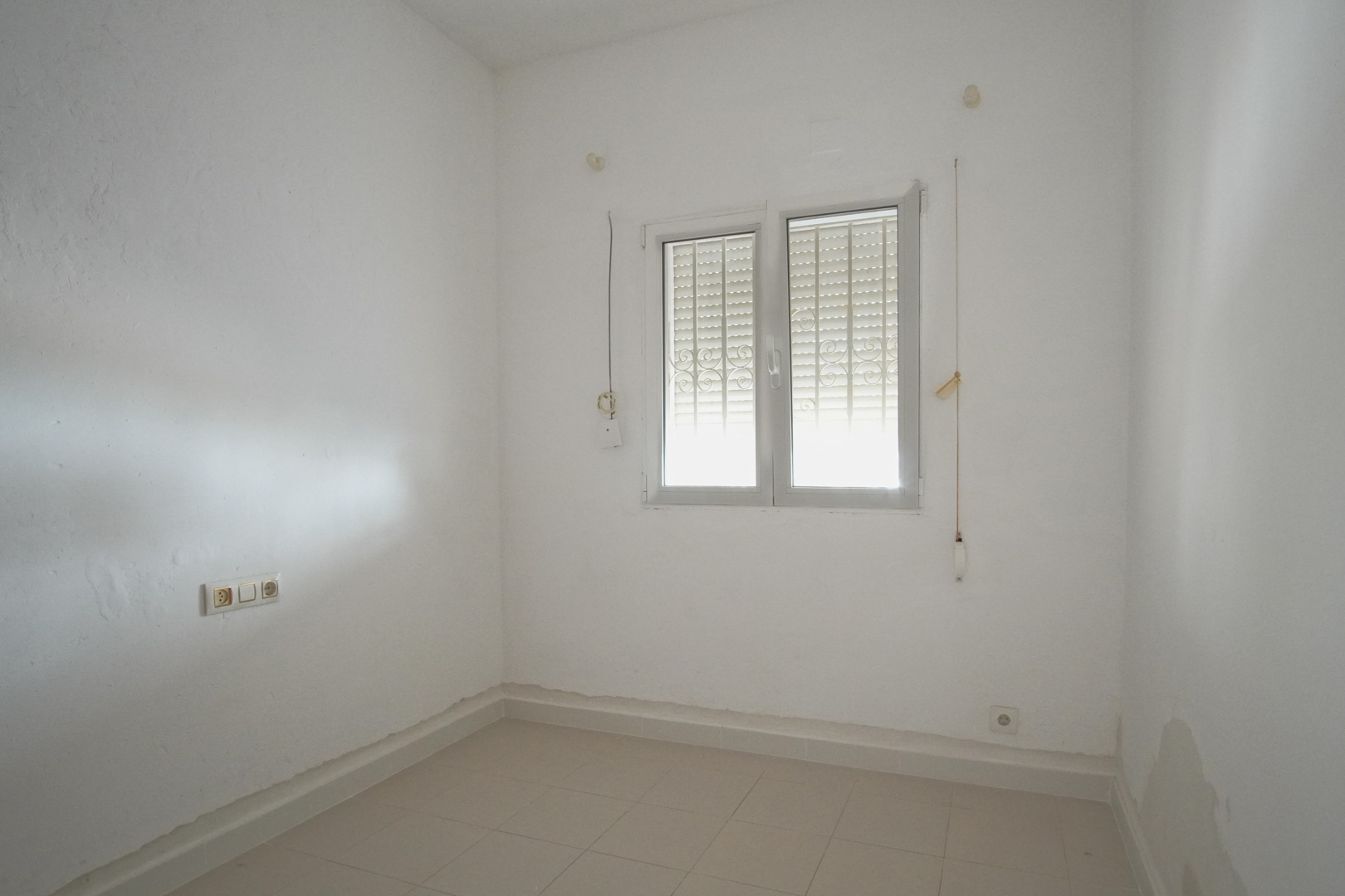 Habitación luminosa con paredes blancas, suelos de gres y ventana de doble hoja con rejas de seguridad y persiana integrada.