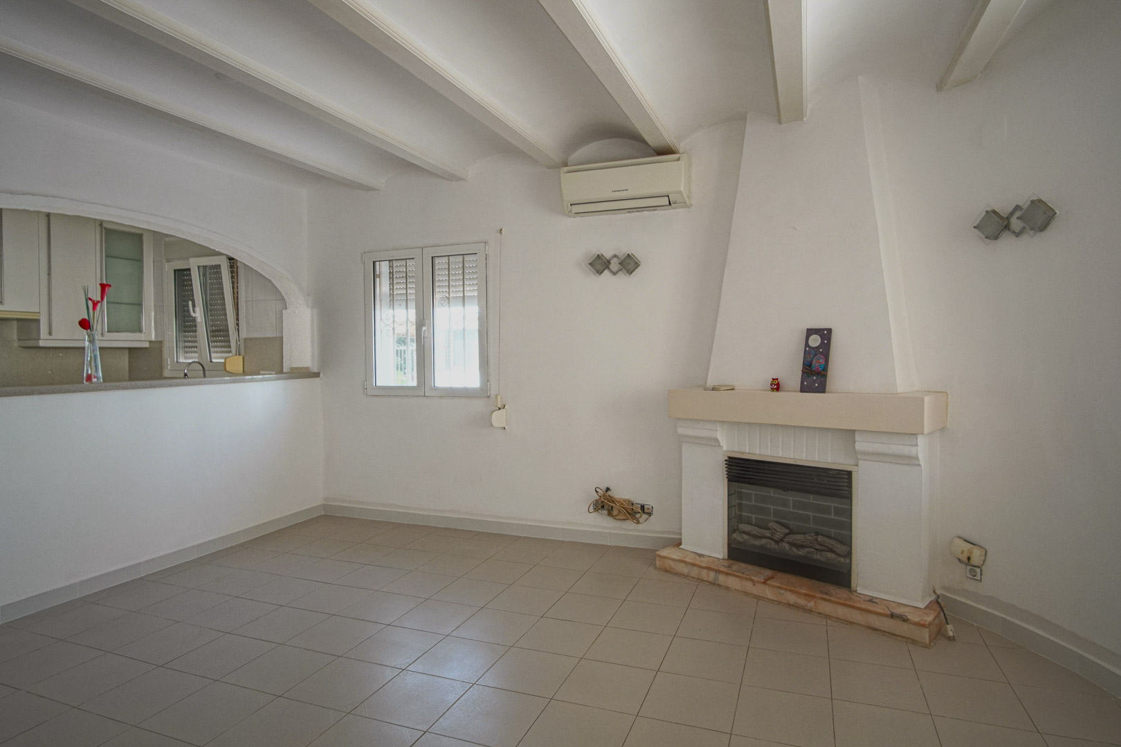 Salón con chimenea de obra, techos de vigas blancas y aire acondicionado. La estancia conecta con la cocina a través de un arco abierto.