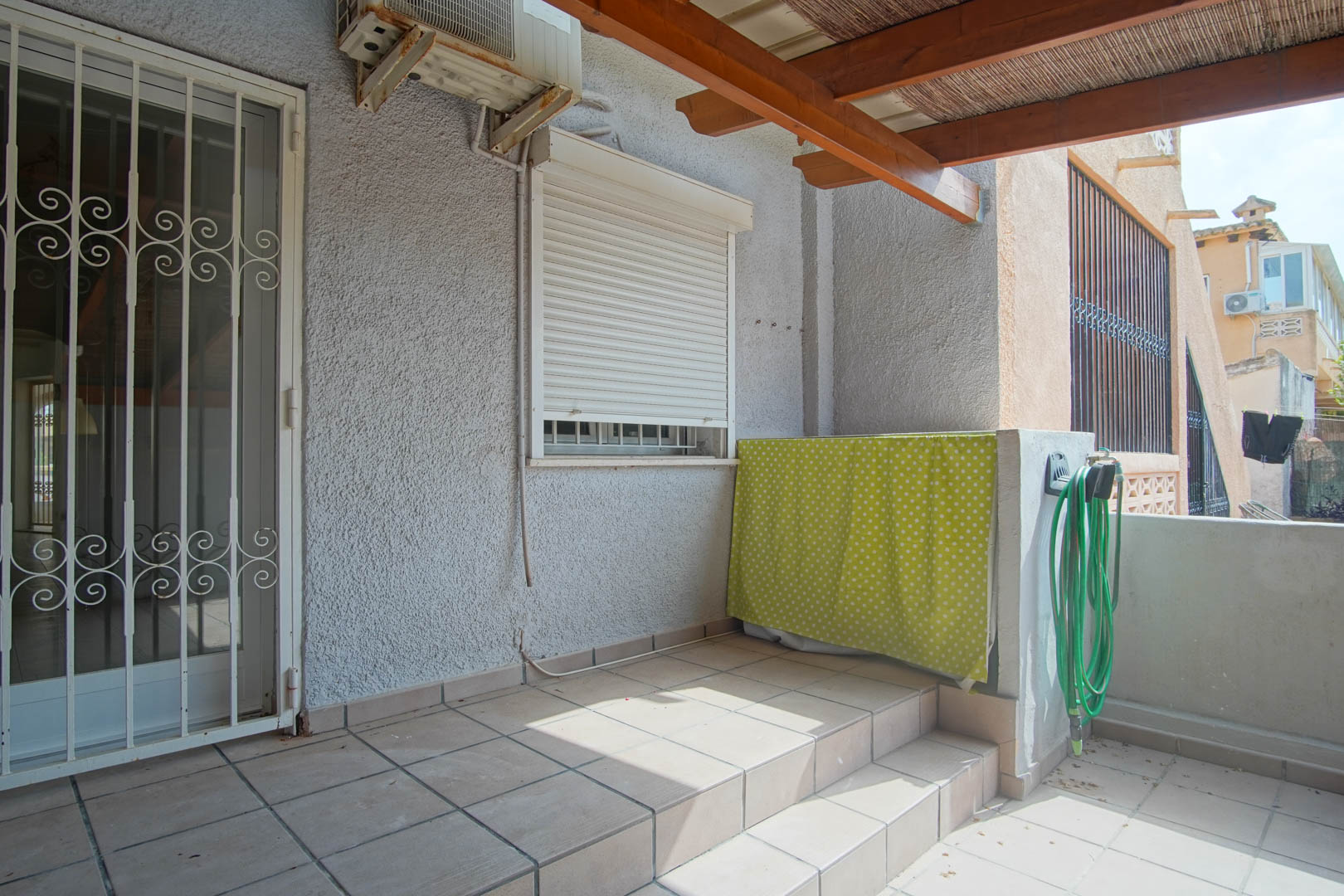 Terraza exterior con suelo cerámico, pérgola de madera con cañizo y acceso protegido por reja de seguridad. Incluye unidad de aire acondicionado y zona de servicio.
