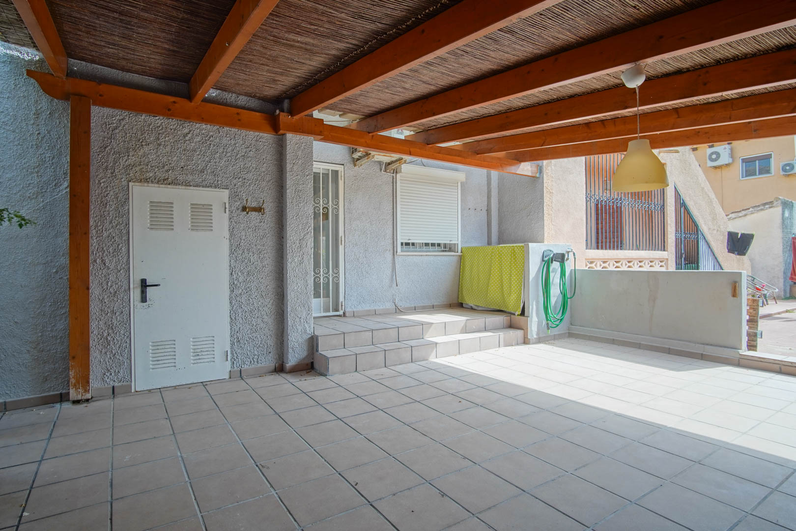 Terraza exterior cubierta con una robusta pérgola de madera y cañizo, suelos cerámicos y acceso elevado a la vivienda mediante escalones y puerta de seguridad blanca.