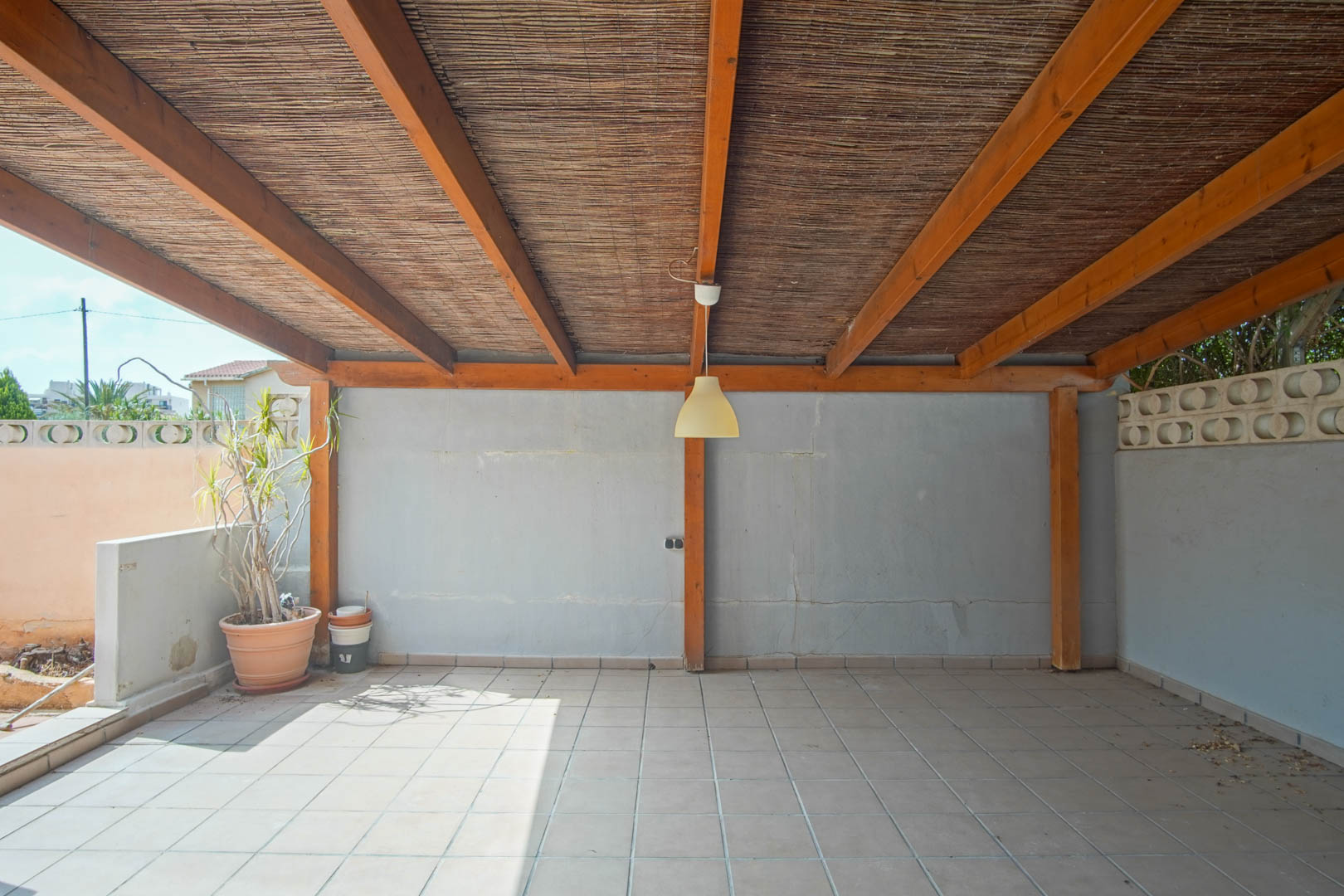 Terraza exterior cubierta con vigas de madera y techado de cañizo, suelo cerámico y muros perimetrales con bloques decorativos.