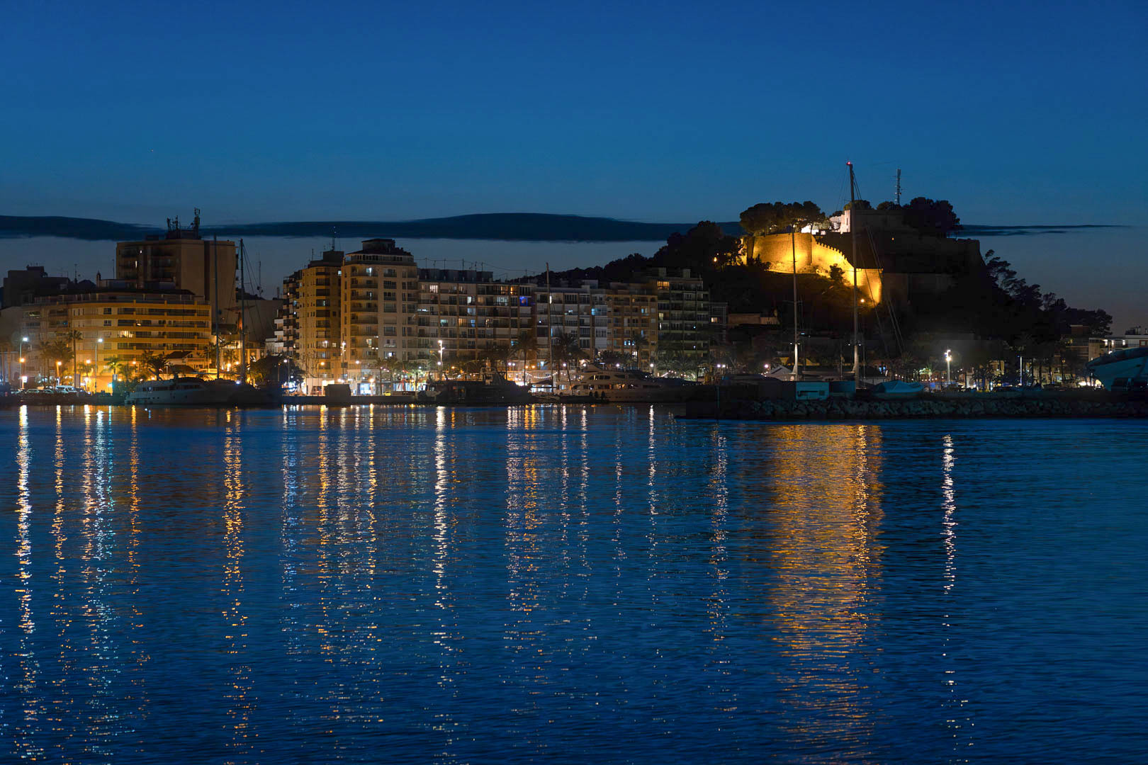 Vista nocturna del puerto de Dénia con el histórico castillo iluminado y el perfil urbano reflejado sobre las tranquilas aguas del Mediterráneo.