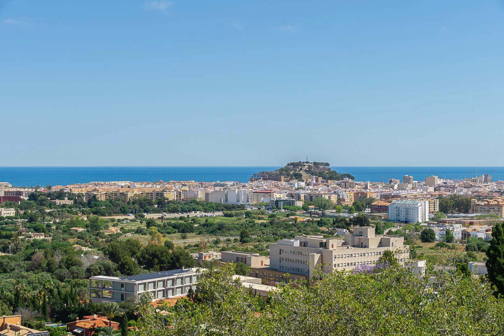 Vista panorámica de Dénia con el castillo histórico sobre la colina, el mar Mediterráneo al fondo y el entorno urbano rodeado de vegetación.