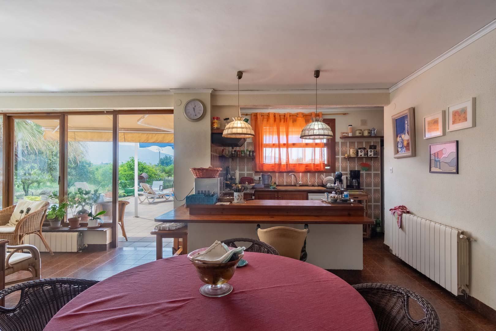 Cocina abierta con barra de madera y comedor integrado, conectada mediante grandes ventanales a una terraza exterior con vistas al jardín mediterráneo.