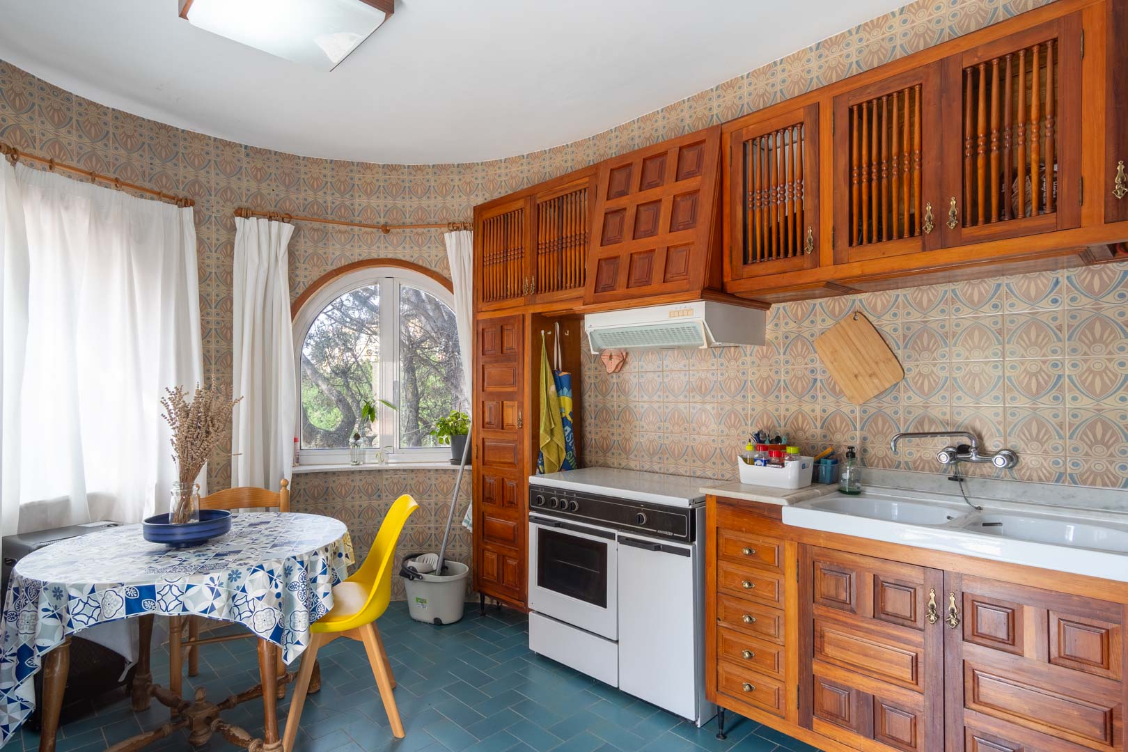Cocina de estilo rústico con mobiliario de madera tallada, azulejos decorativos y una zona de desayuno junto a un ventanal de arco con vistas al jardín.
