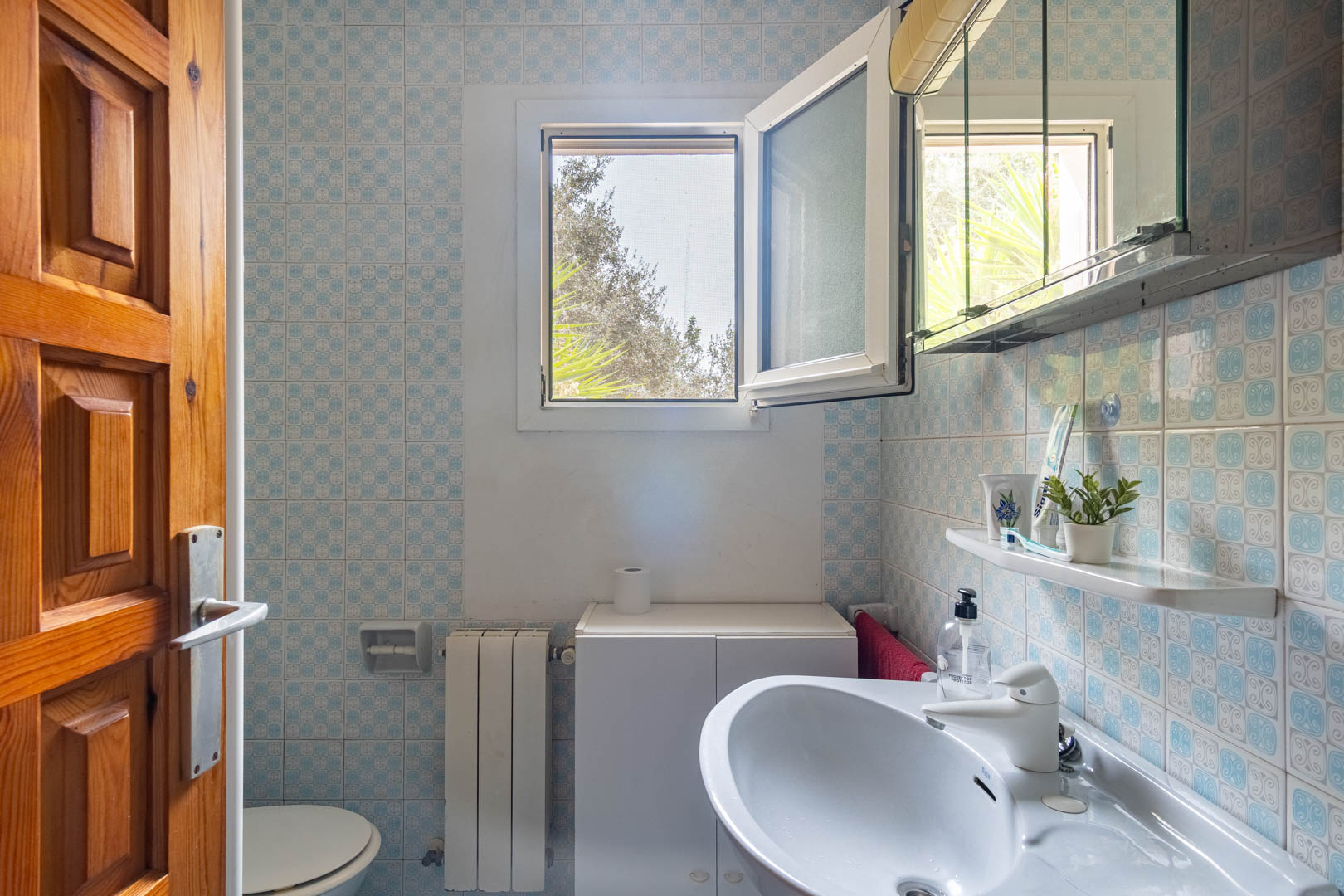 Cuarto de baño con azulejos decorativos en tonos azules, lavabo blanco de cerámica y ventana oscilobatiente que aporta luz natural y ventilación.