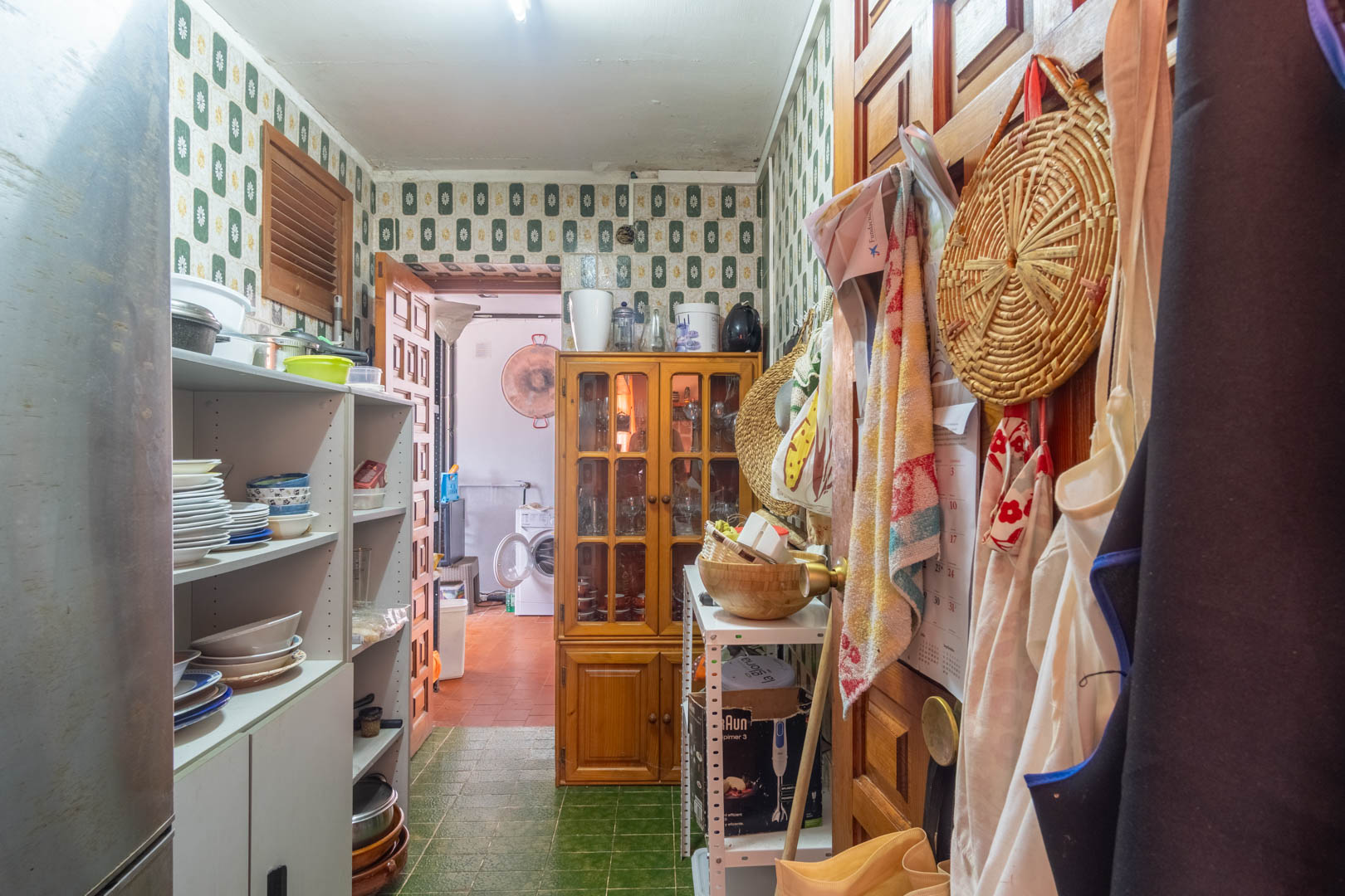 Despensa funcional con azulejos de época, estanterías para vajilla y una vitrina de madera, conectada directamente con la zona de lavandería.