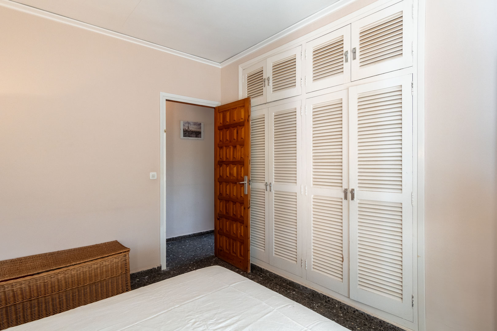 Dormitorio con amplio armario empotrado de puertas mallorquinas blancas, suelo de terrazo oscuro y puerta de madera maciza tallada que da acceso al pasillo.