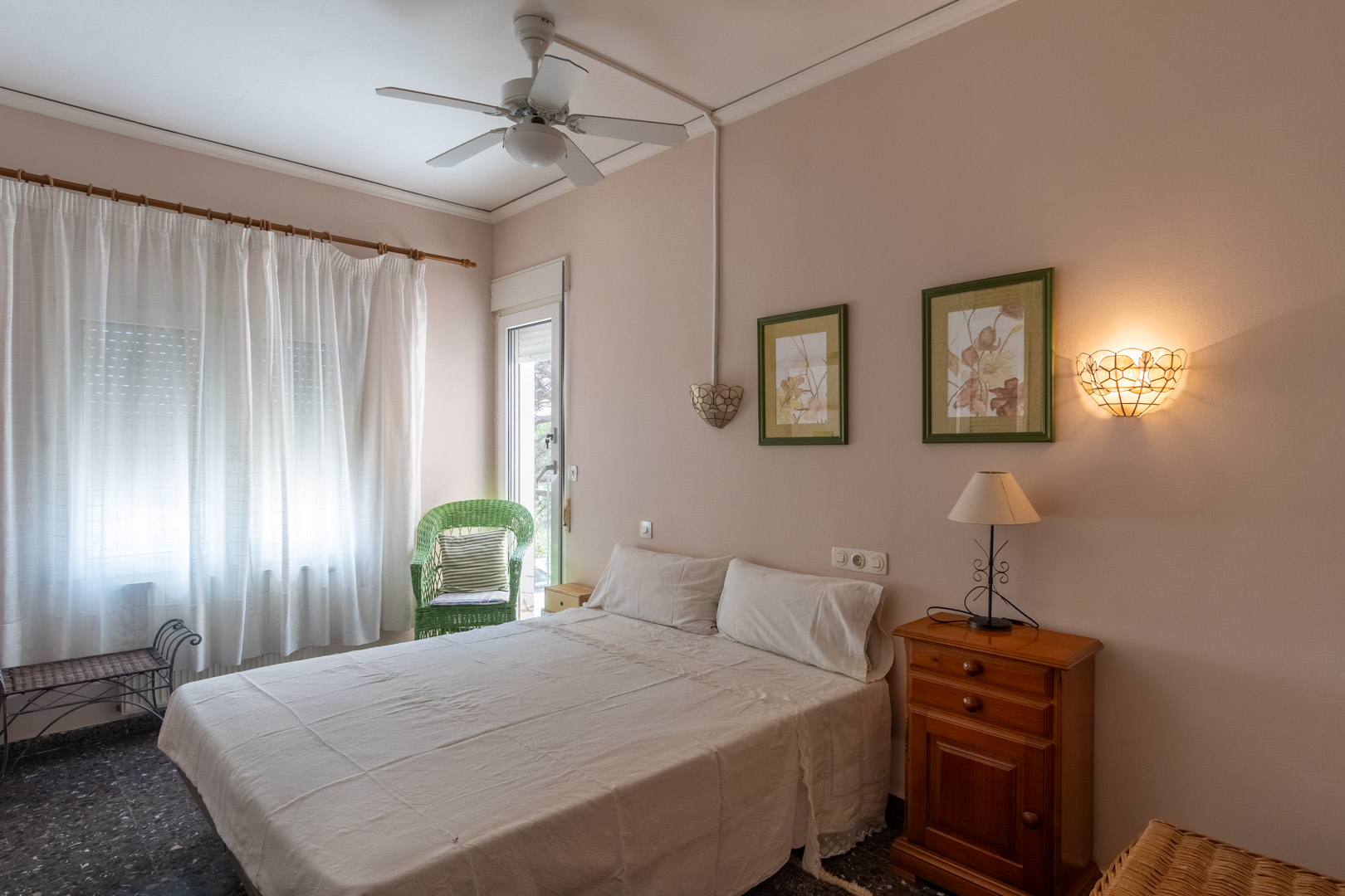 Dormitorio con cama doble, ventilador de techo y acceso directo al exterior. Incluye mobiliario de madera, suelos de terrazo y una silla de mimbre decorativa junto al ventanal.