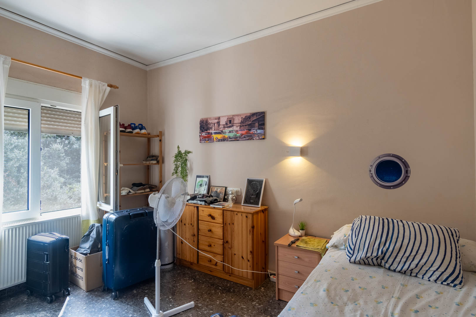 Dormitorio con suelos de terrazo, mobiliario de madera natural y ventana al exterior que aporta luz natural. Incluye calefacción por radiador y aplique de pared.