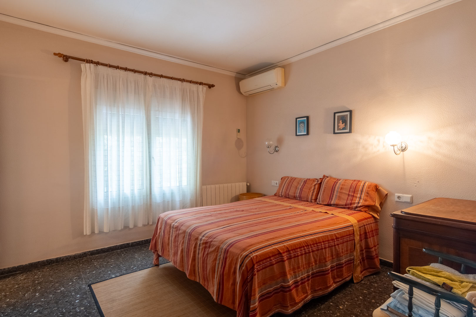 Dormitorio doble con suelos de terrazo oscuro, equipado con aire acondicionado, radiador y ventana con cortinas blancas que filtran la luz natural.