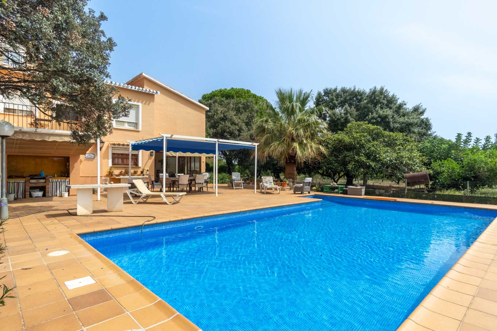 Piscina rectangular con revestimiento de mosaico azul, rodeada de una amplia terraza de gres y zona de barbacoa junto a la vivienda de estilo mediterráneo.