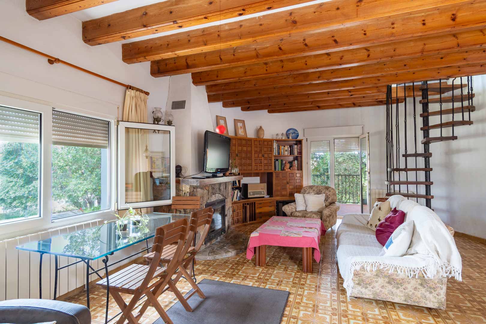 Salón comedor con techos de vigas de madera vistas, chimenea de piedra y una escalera de caracol metálica que conecta con la planta superior.