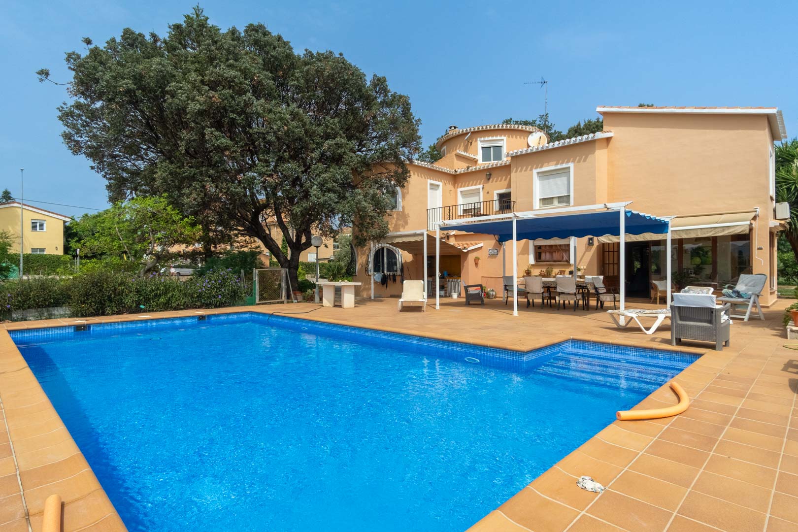 Vista exterior de la piscina rectangular rodeada de una amplia terraza de gres, con la fachada mediterránea y una zona de comedor bajo toldo azul al fondo.