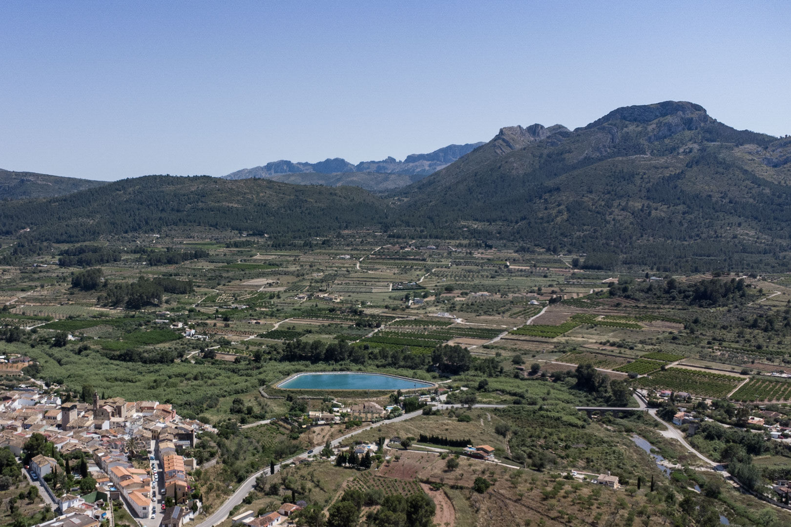 Vista aérea panorámica del valle de Jalón desde Alcalalí, mostrando el entorno natural, parcelas agrícolas, un embalse turquesa y las montañas circundantes.