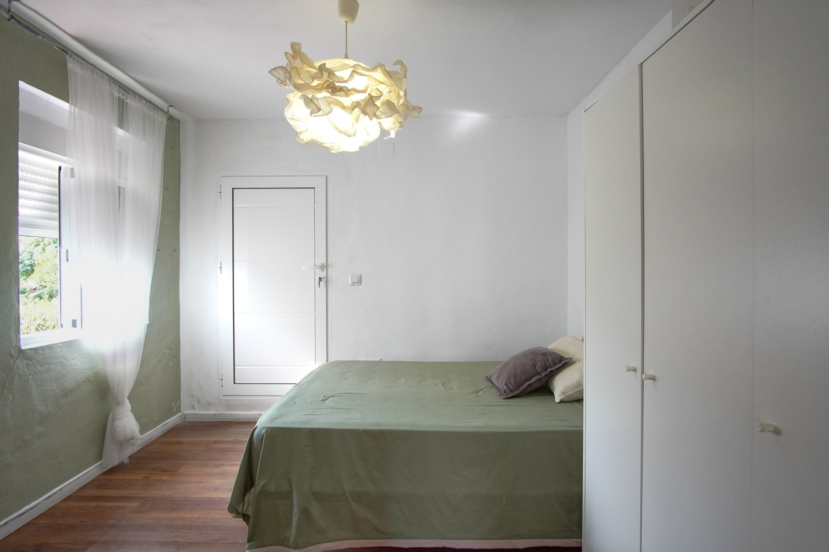 Habitación luminosa con cama cubierta de lino verde, lámpara de diseño y armario empotrado blanco.