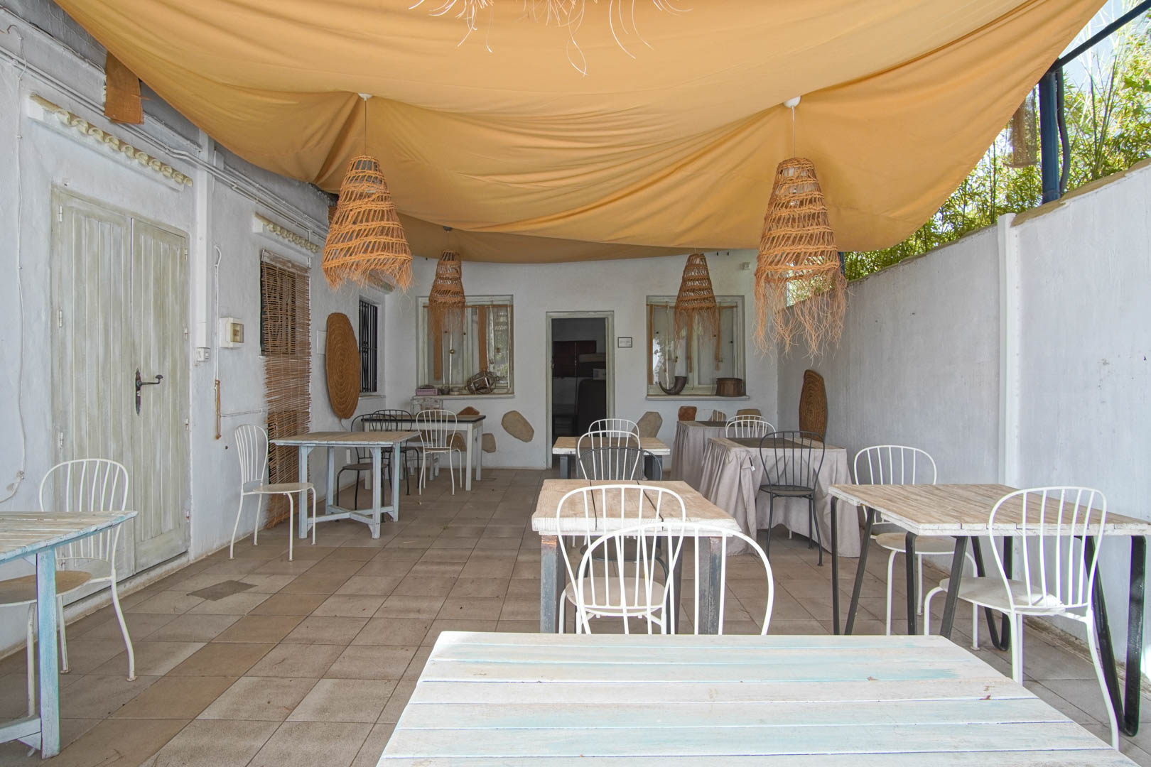 Porche de restaurante con mesas de madera y lámparas colgantes de mimbre bajo toldo.