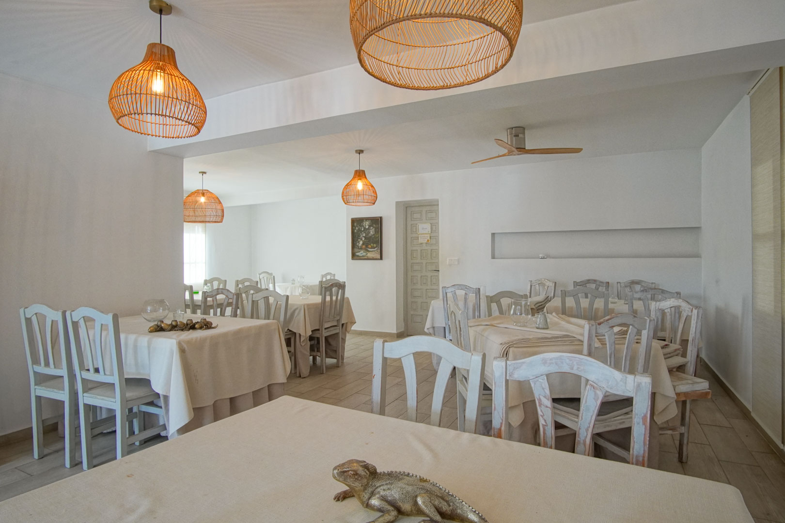 Salón de restaurante luminoso con mesas vestidas y sillas de madera pintadas.