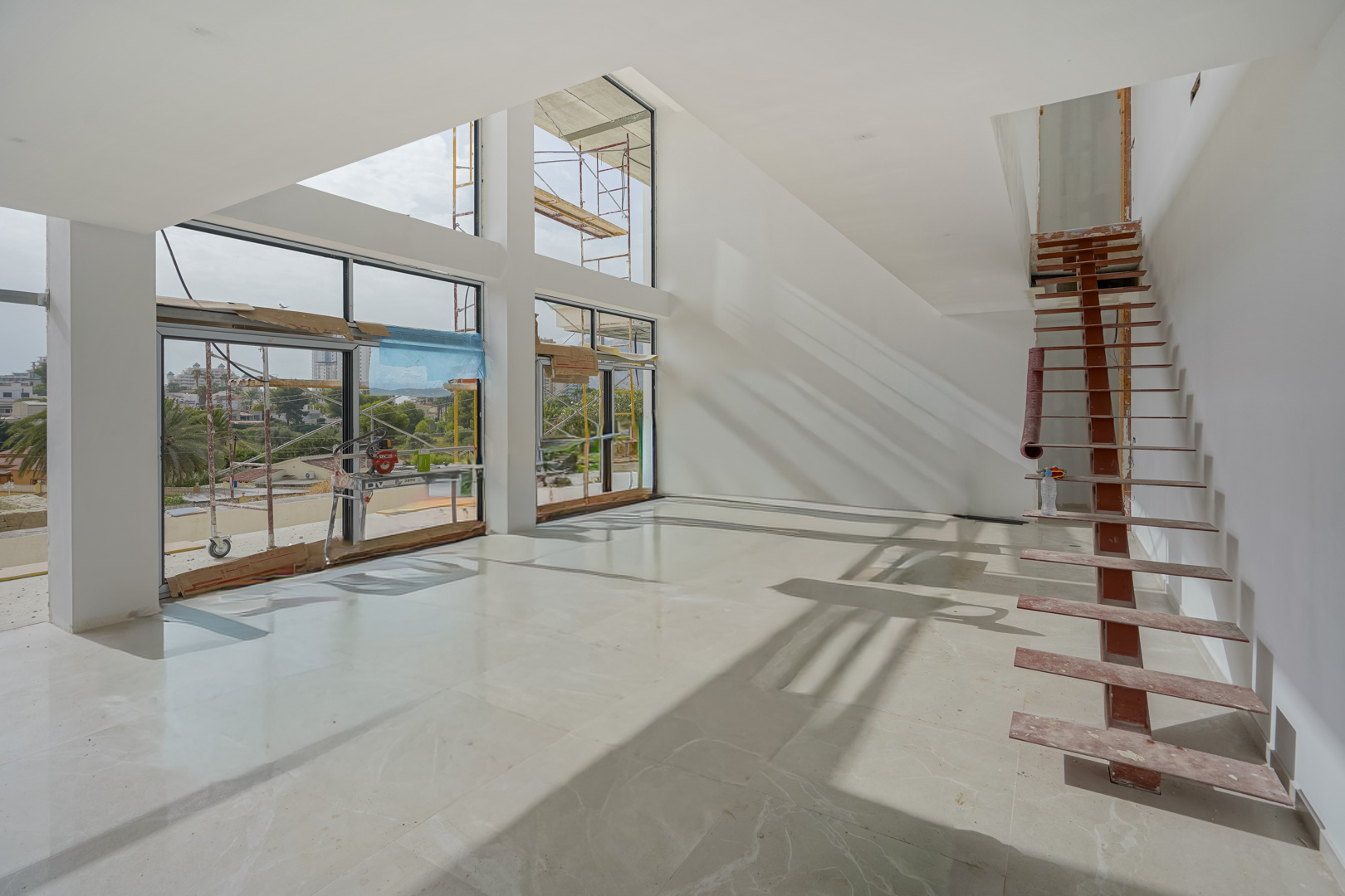 Salón de doble altura en construcción con amplios ventanales, suelos de piedra pulida y estructura de escalera metálica. La luz natural inunda el espacio diáfano.