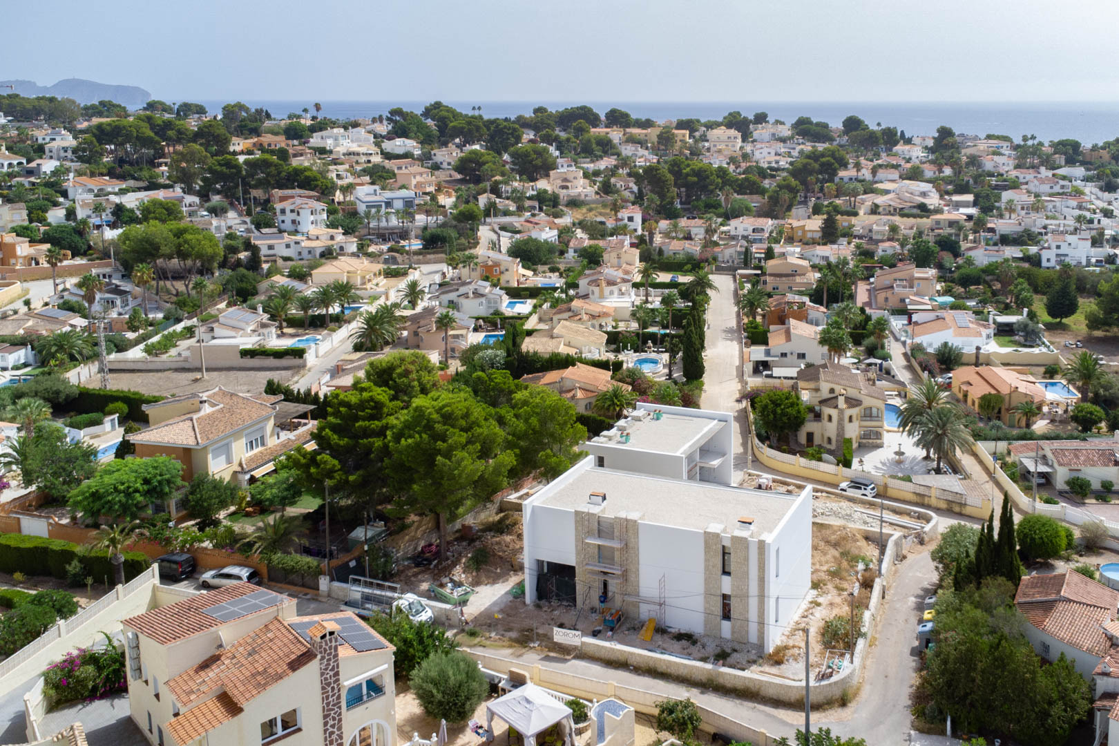 Vista aérea de la villa en construcción con fachada blanca y detalles en piedra natural, ubicada en una zona residencial de Calpe con el mar en el horizonte.
