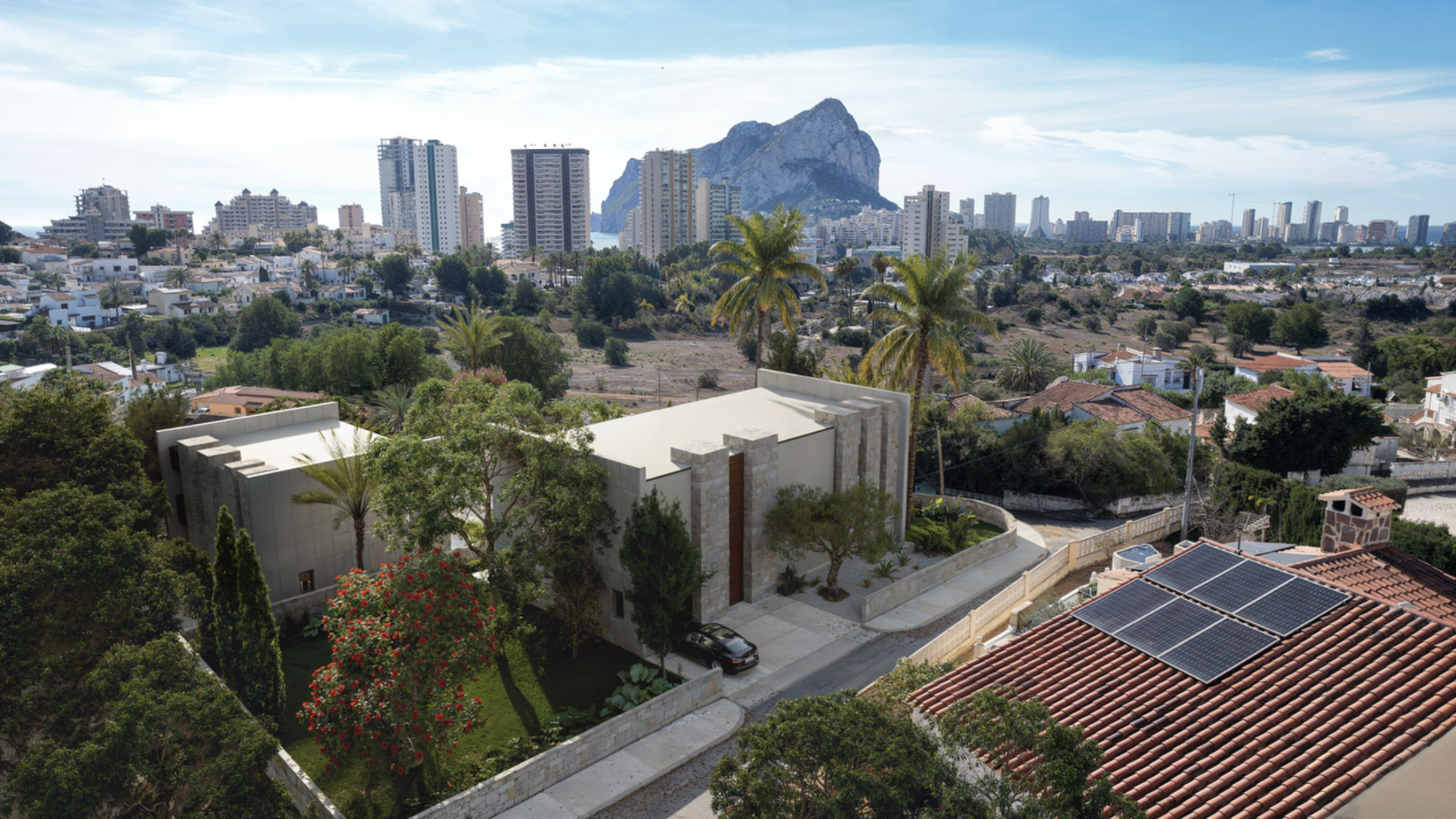 Vista aérea de la villa moderna con fachada de piedra natural y jardín, destacando las vistas panorámicas al Peñón de Ifach y el skyline de Calpe.