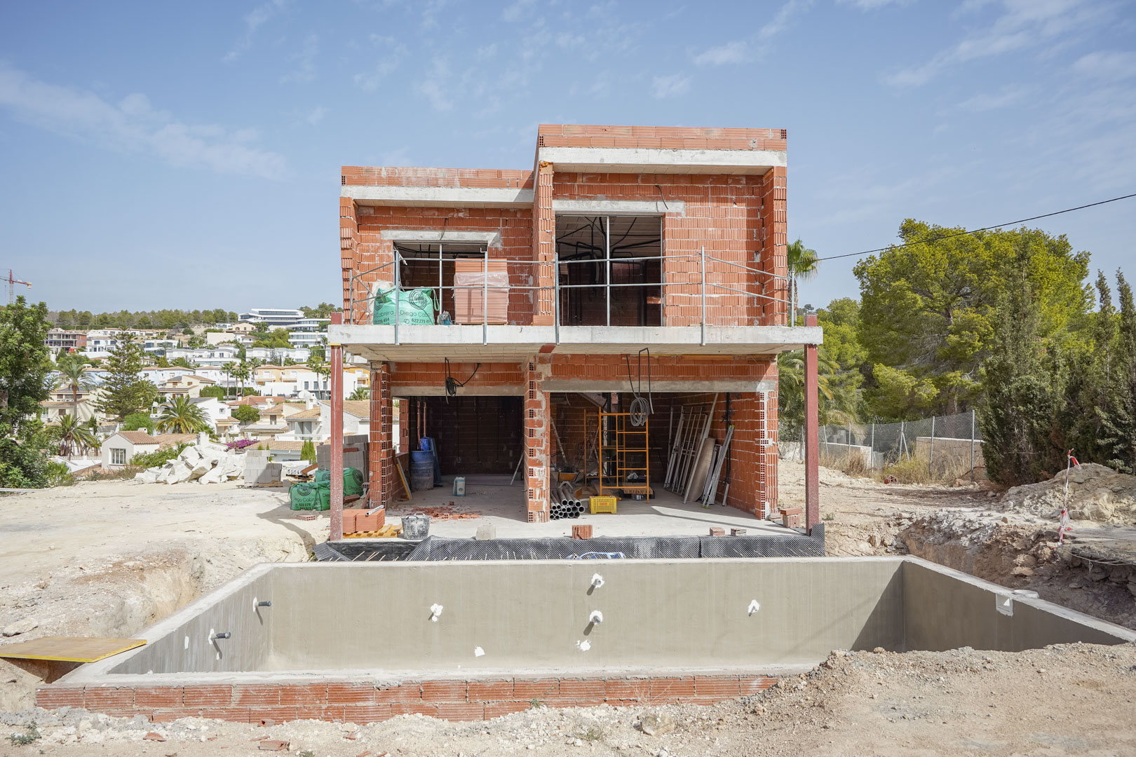 Estado de obra de la villa con estructura de ladrillo de dos plantas y el vaso de la piscina de hormigón en primer plano bajo un cielo despejado.