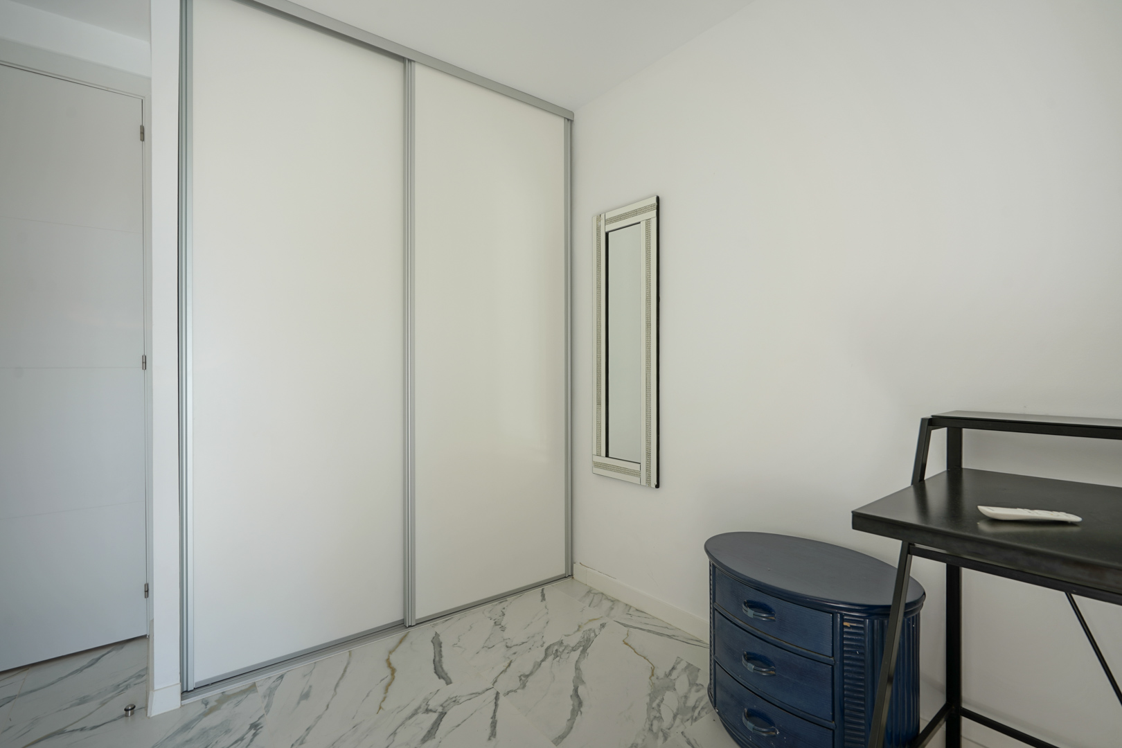 Armario empotrado con puertas correderas blancas sobre suelo de mármol veteado, complementado con un espejo vertical y mobiliario auxiliar.