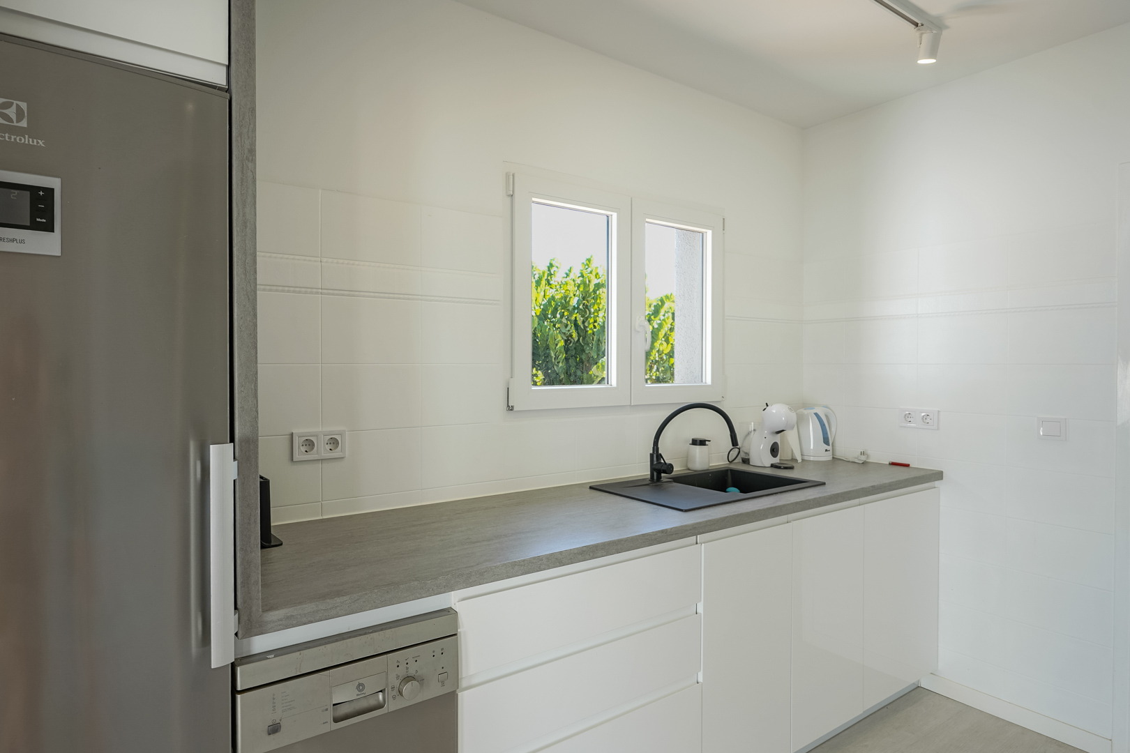 Cocina funcional con armarios blancos sin tiradores, encimera gris, fregadero negro y electrodomésticos integrados bajo una ventana con luz natural.