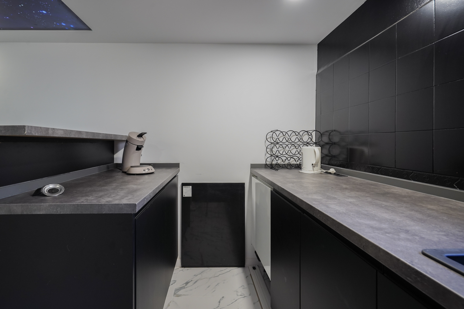 Cocina secundaria con armarios negros, encimeras grises y suelo de mármol. Incluye un panel de techo decorativo con efecto de cielo estrellado.