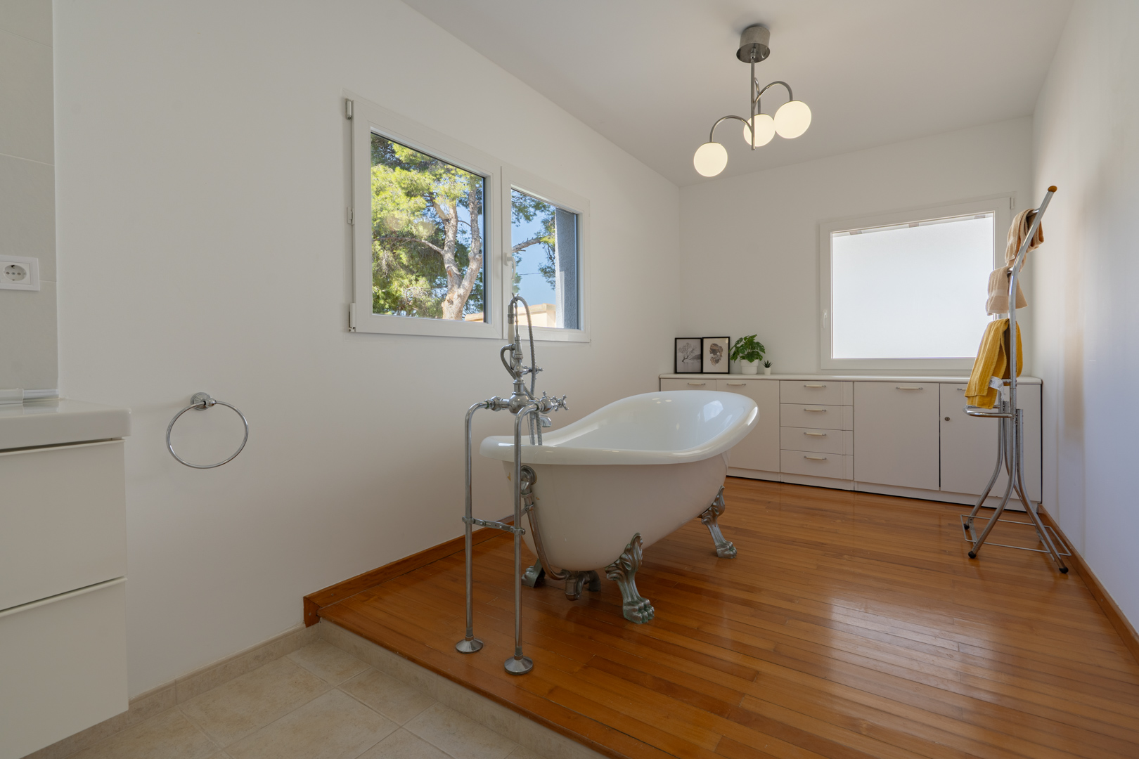 Cuarto de baño con bañera exenta de patas clásicas sobre tarima de madera, mobiliario blanco integrado y ventanas de doble acristalamiento con vistas al exterior.