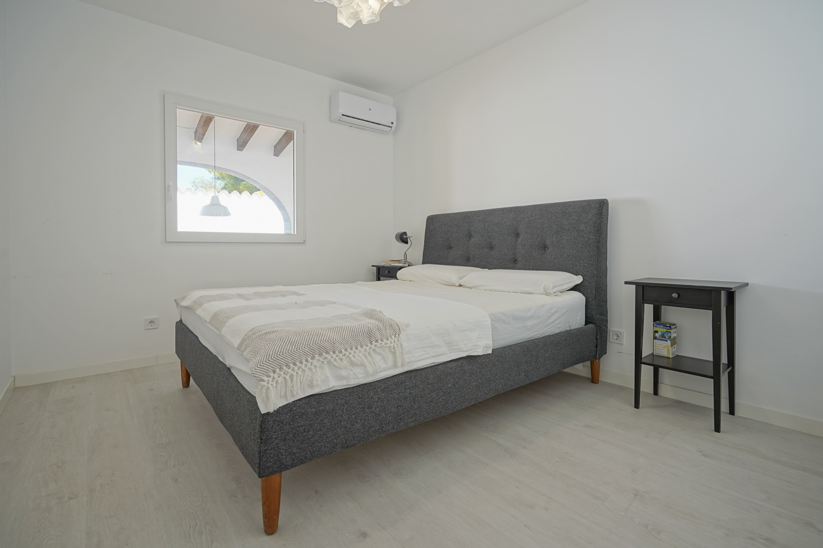 Dormitorio luminoso con cama doble tapizada en gris, aire acondicionado y suelos de madera clara. La ventana ofrece vistas a la terraza cubierta con vigas de madera.